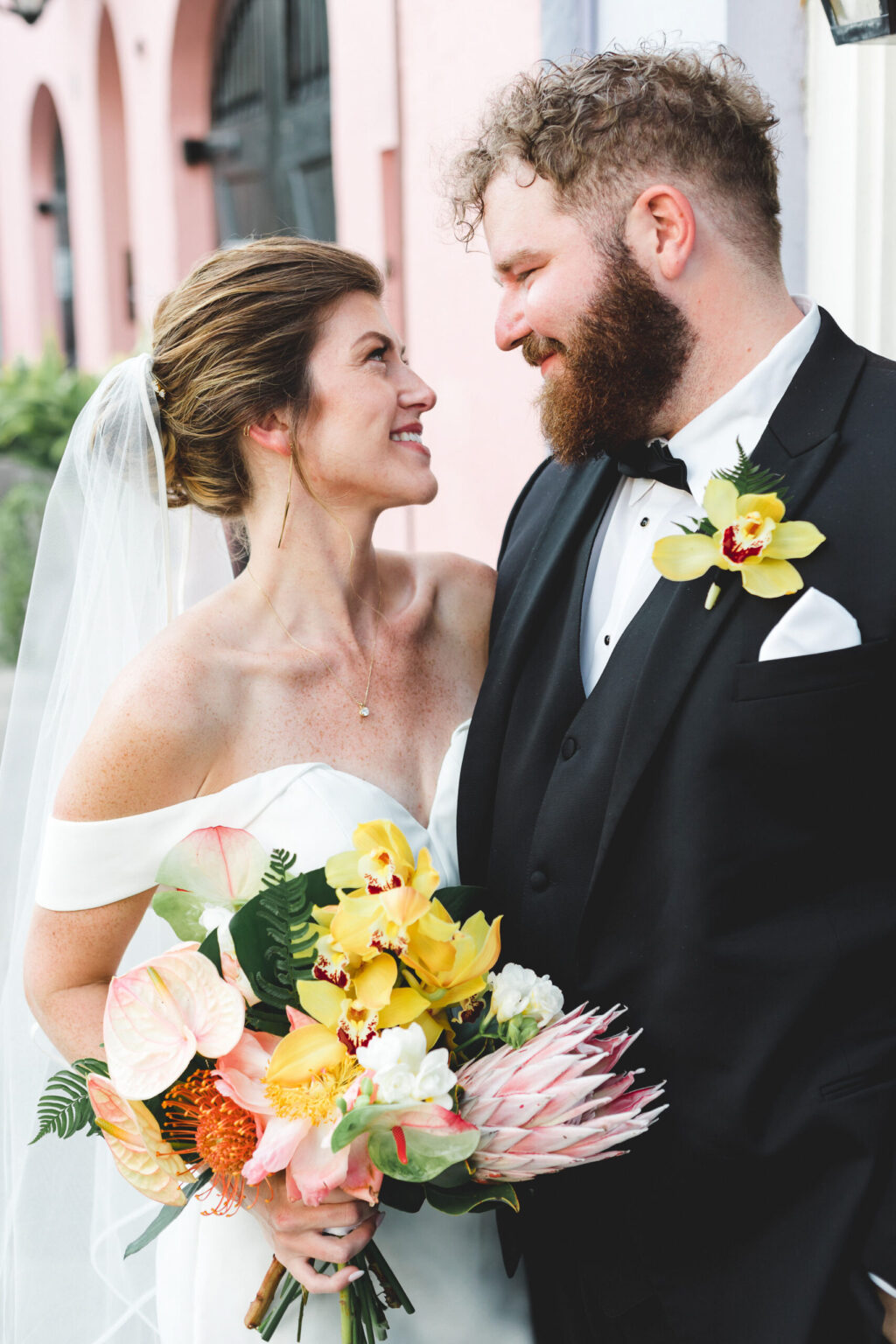 Elopement Wedding A bride and groom gaze lovingly at each other on their wedding day. Having chosen to elope, the bride is dressed in an off-the-shoulder white gown and holds a vibrant bouquet of yellow, pink, and white flowers. The groom, wearing a black tuxedo with a yellow orchid boutonniere, stands close to her. Elopements Inc