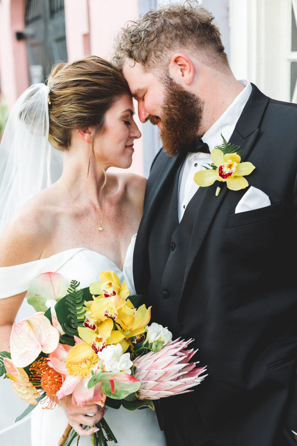 Elopement Wedding A newly married couple, who decided on a romantic elopement, leans in with their foreheads touching, smiling affectionately. The bride, wearing an off-shoulder white gown and veil, holds a vibrant bouquet of yellow and pink flowers. The groom, in a black tuxedo adorned with a yellow orchid boutonniere, gazes lovingly at her. Elopements Inc