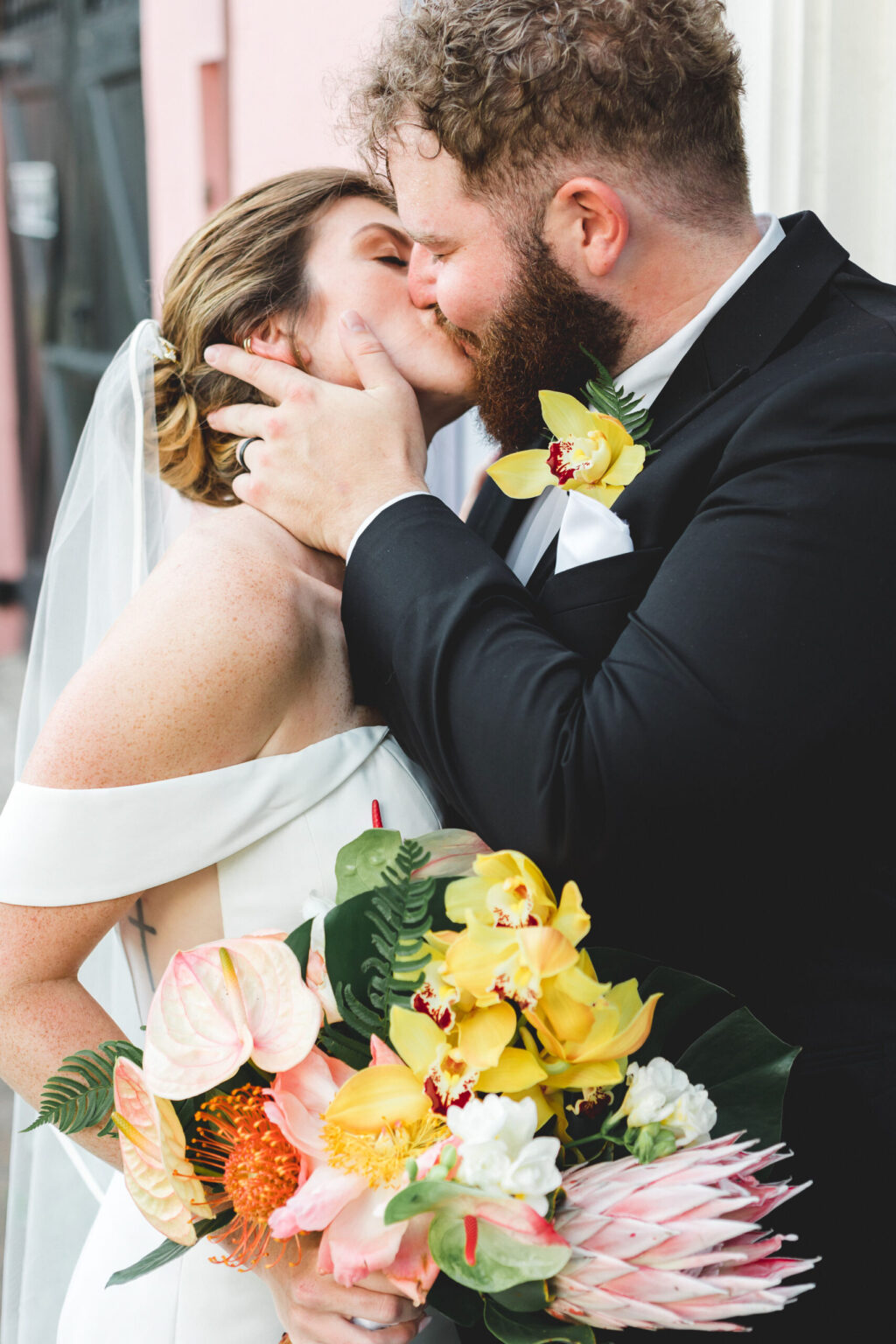 Elopement Wedding A bride and groom share a kiss on their elopement. The bride, wearing an off-shoulder white gown and veil, holds a vibrant bouquet of flowers, including yellow orchids and pink anthuriums. The groom, in a black suit with a yellow orchid boutonniere, lovingly embraces her face. Elopements Inc