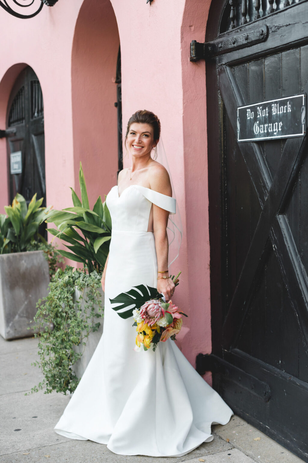 Elopement Wedding A bride in a white off-the-shoulder mermaid wedding dress holds a tropical bouquet while standing against a pink wall with black arched doors. A sign on the door reads "Do Not Block Garage." She smiles, one hand on her hip near a large planter with verdant plants, ready to elope. Elopements Inc