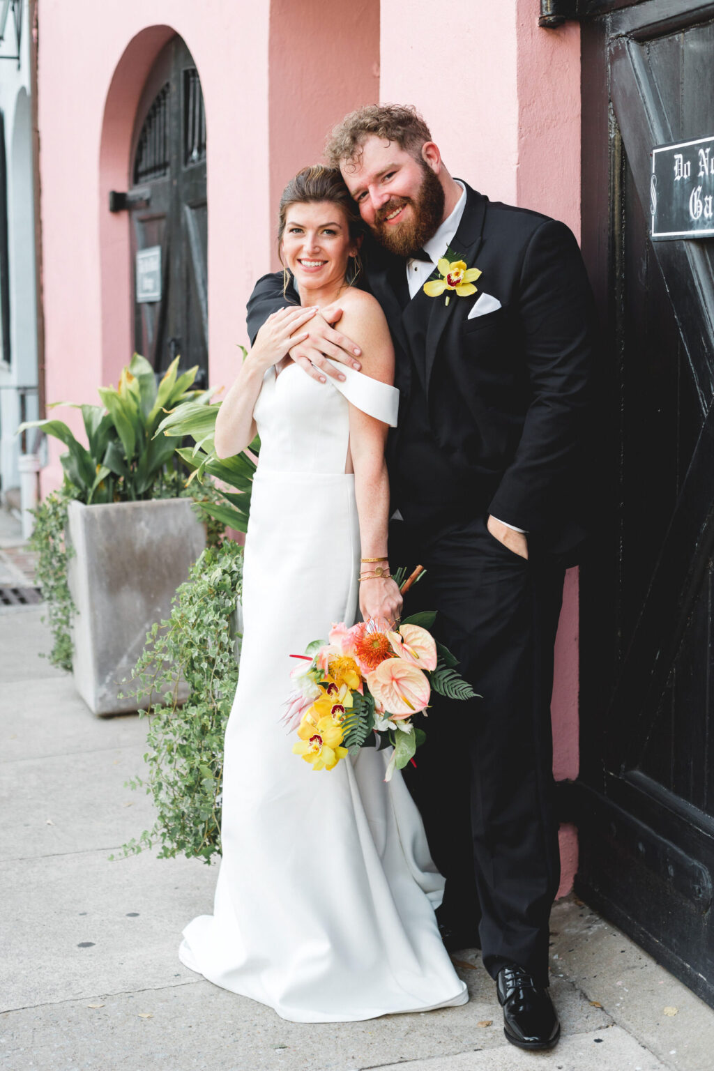 Elopement Wedding Bride in an off-shoulder white gown with a floral bouquet and groom in a black tuxedo with a yellow boutonniere pose joyfully against a pink building. The groom, with a full beard, leans against the wall, smiling, while the bride stands beside him, her hand resting on his chest—capturing their intimate elopement moment perfectly. Elopements Inc