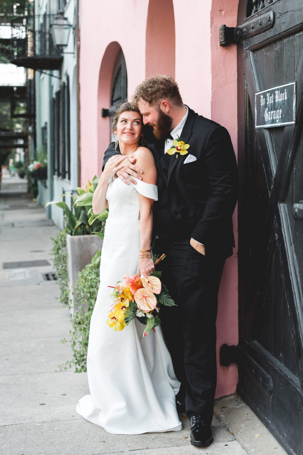 Elopement Wedding A bride in a white dress holds a colorful bouquet and smiles while leaning against a groom in a black suit. They stand in front of a pink wall next to a wooden door with a sign reading "Do Not Block Garage." Their intimate elopement is filled with joy and warmth, lush potted plants, and a distant building in the background. Elopements Inc