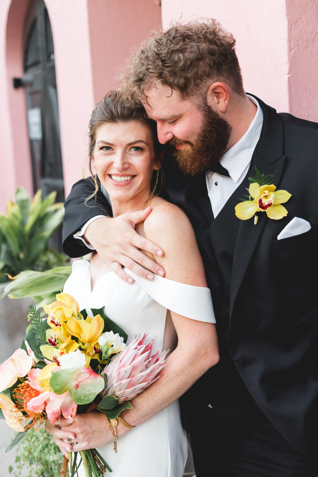 Elopement Wedding A bride and groom smile happily while standing outside, celebrating their elopement. The bride, in a white off-shoulder dress, holds a vibrant bouquet of flowers. The groom, wearing a black suit with a yellow boutonniere, has his arm affectionately around the bride's shoulders. The background features a pink building and greenery. Elopements Inc