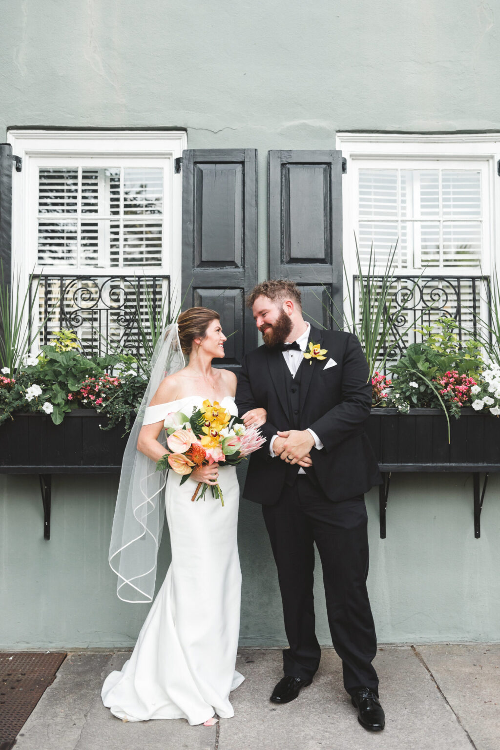 Elopement Wedding A bride in an off-shoulder white gown holds a colorful bouquet, standing beside a groom in a black tuxedo. They are smiling at each other, having just eloped, in front of a light green wall with two black-shuttered windows adorned with flower boxes containing white and pink flowers. Elopements Inc