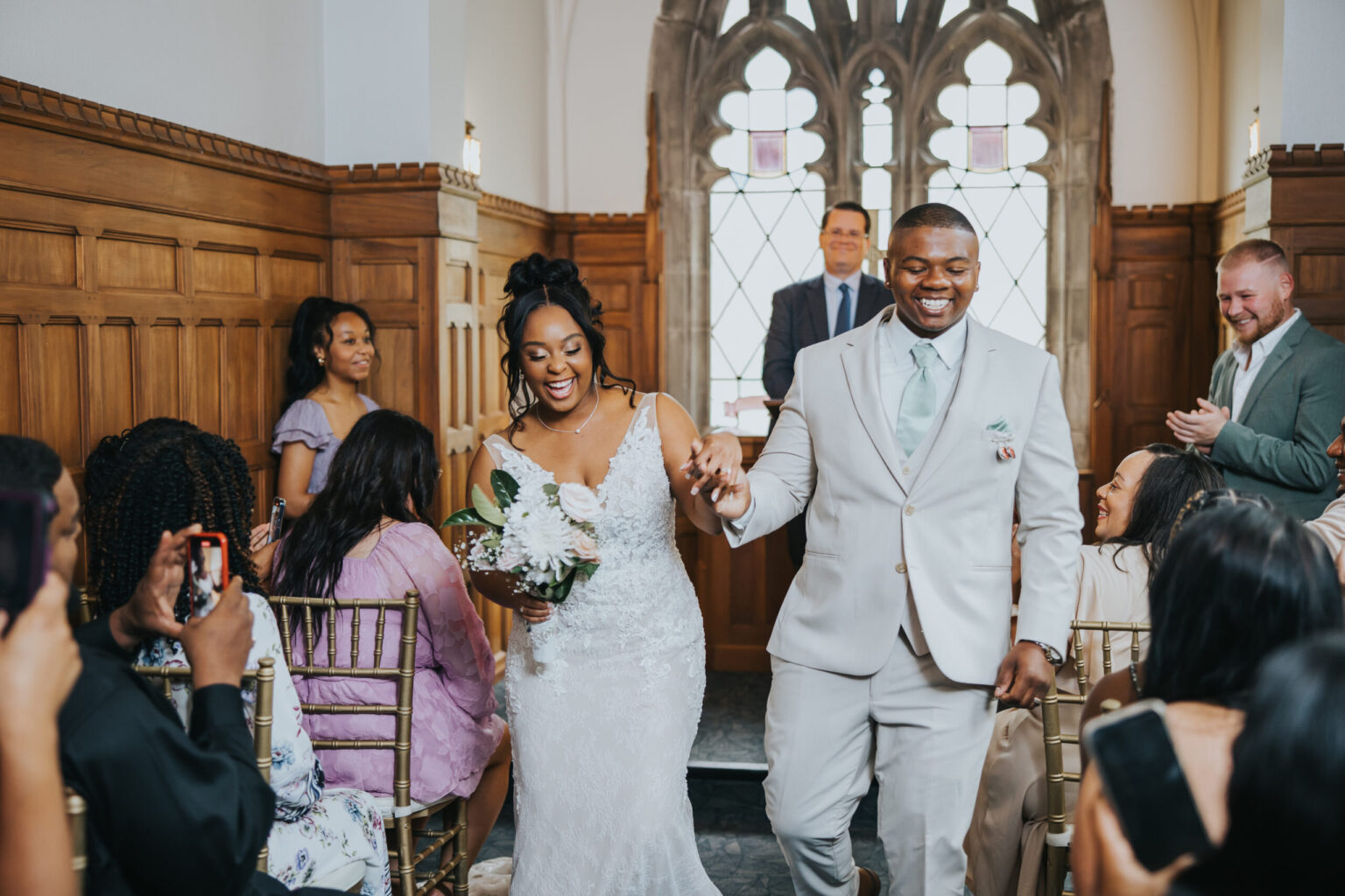 Elopement Wedding A bride in a lace wedding gown and a groom in a light-colored suit, holding hands and smiling, walk down the aisle of a wood-paneled room after their intimate elopement ceremony. Guests seated on both sides, many capturing the moment with their phones. A minister smiles in the background. Elopements Inc