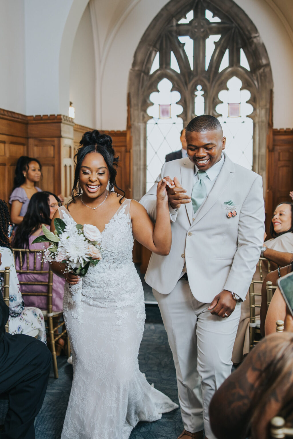 Elopement Wedding A joyful couple walks down the aisle inside a church after their intimate elopement. The bride wears a white lace gown and holds a bouquet, while the groom sports a light gray suit, holding her hand. Guests sit on either side, watching and smiling. Sunlight filters through the arched windows behind them. Elopements Inc