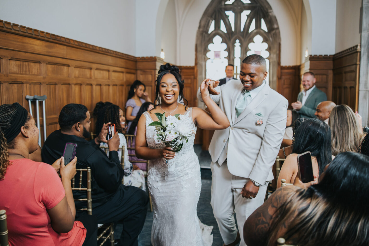 Elopement Wedding A newlywed couple, dressed in elegant wedding attire, joyfully walks down the aisle of a charming church with arched doorways and wooden paneling. The bride holds a bouquet, and the groom raises their hands together. Guests, seated on both sides, smile and capture the moment on their phones; one can't help but think they might have eloped to create such an intimate celebration. Elopements Inc