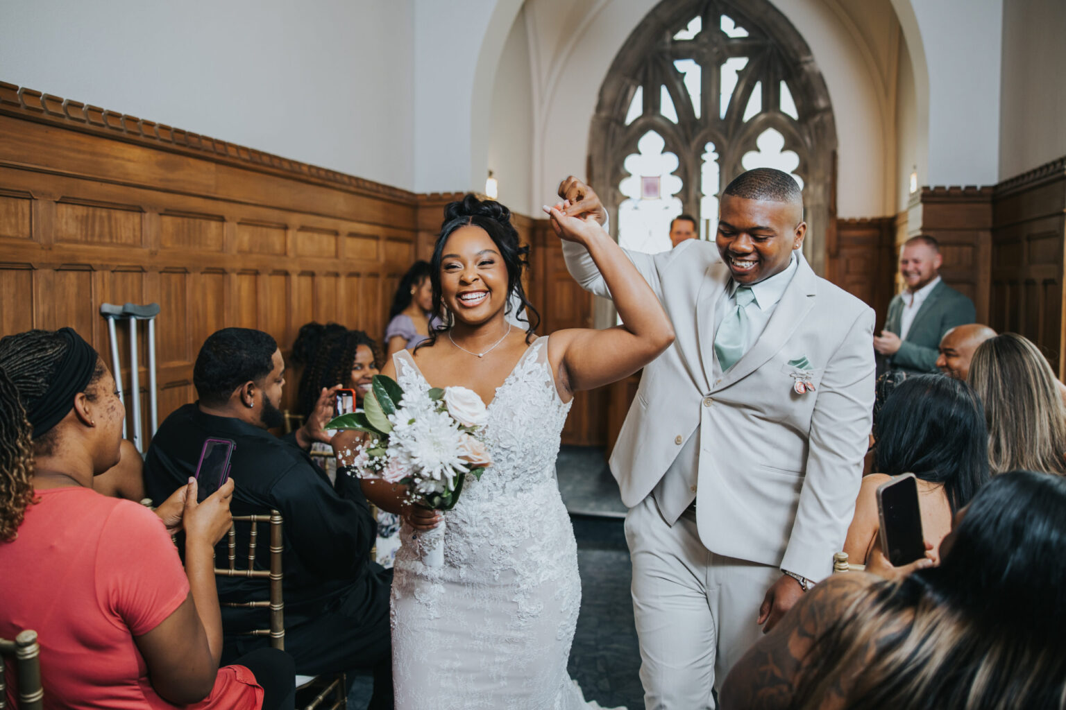 Elopement Wedding A joyful bride and groom walk hand in hand down the aisle. The bride wears a white lace gown and holds a bouquet of white flowers, while the groom dons a light gray suit with a mint green tie. Guests seated on either side cheer and take photos in a beautifully decorated, wood-paneled venue celebrating their elopement. Elopements Inc