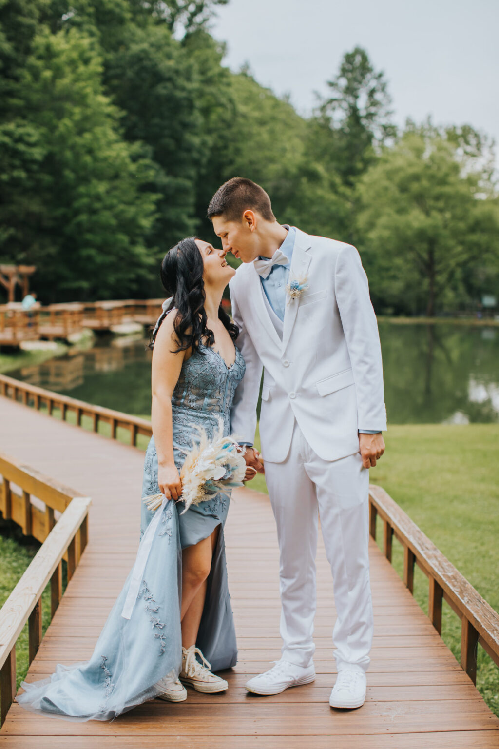 Elopement Wedding A couple stands on a wooden walkway by a lush, green park with a pond in the background. They are dressed in formal attire; the woman wears a blue dress with a train and the man is in a white suit with a blue bow tie. They lean in for an intimate kiss, holding hands and wearing white sneakers, celebrating their elopement. Elopements Inc