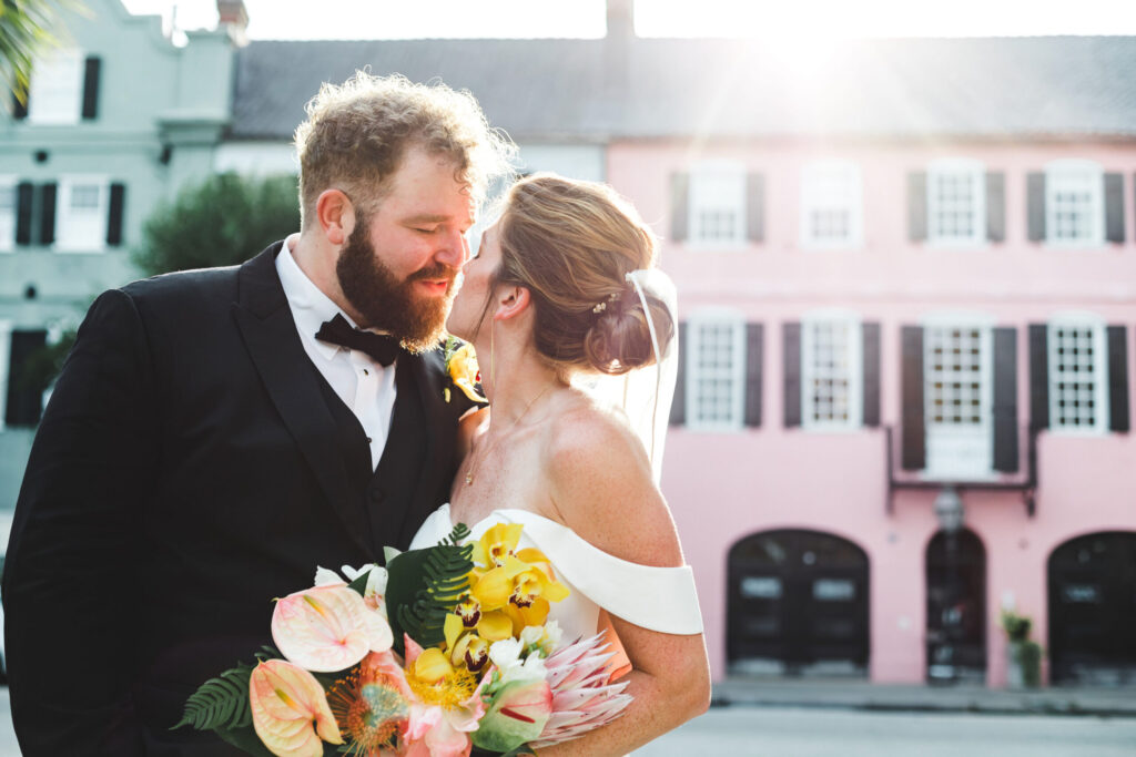 Elopement Wedding A groom in a black tuxedo and bowtie and a bride in an off-the-shoulder white wedding gown with a veil share a close, joyful moment while holding a vibrant bouquet of flowers. They stand in front of colorful buildings, with sunlight creating a warm, romantic atmosphere perfect for eloping. Elopements Inc