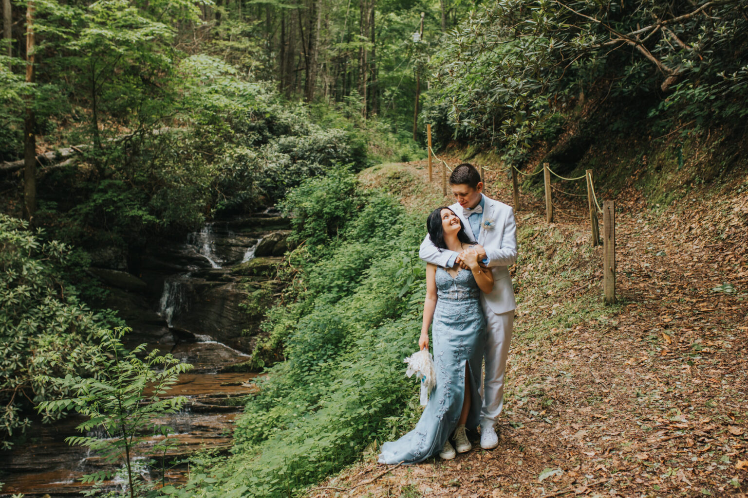 Elopement Wedding A couple stands affectionately in a lush, forested area beside a small waterfall. The man, wearing a light grey suit, hugs the woman in a light blue dress from behind. A narrow, rope-lined path runs alongside the greenery. Both are smiling, enjoying their elopement in the serene natural setting. Elopements Inc