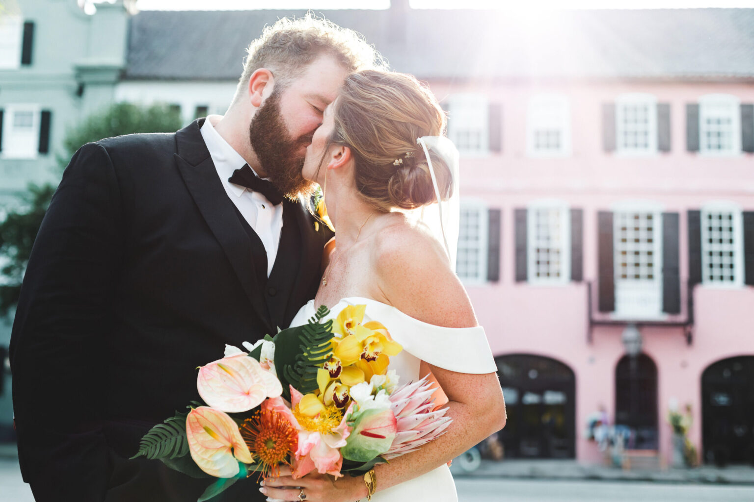 Elopement Wedding A bride and groom share a kiss outdoors during the day. The groom wears a black suit and bowtie, while the bride dons a white off-shoulder dress and veil, holding a colorful bouquet with pink, yellow, and orange flowers. They stand in front of a pastel-colored building with large windows, celebrating their intimate elopement. Elopements Inc