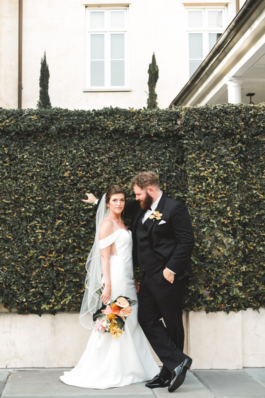 Elopement Wedding A bride in a white gown with a veil and a groom in a black suit stand in front of a lush green ivy wall, having just eloped. The bride holds a bouquet of flowers as the groom leans casually against the wall, both smiling gently, set against the backdrop of an elegant building with tall windows. Elopements Inc