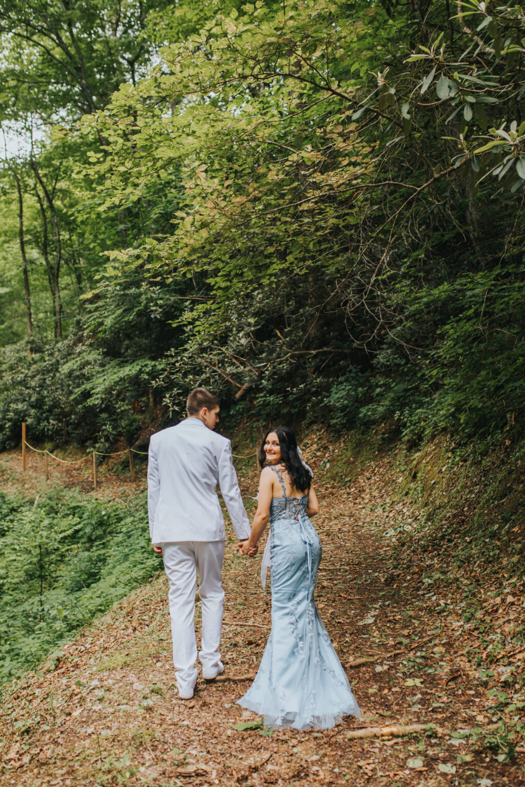 Elopement Wedding A couple, with the woman in a light blue gown and the man in a light gray suit, walk hand in hand along a forest trail. Surrounded by lush greenery and trees, their elopement creates a romantic and serene atmosphere. They glance back, smiling, as they continue their peaceful stroll. Elopements Inc