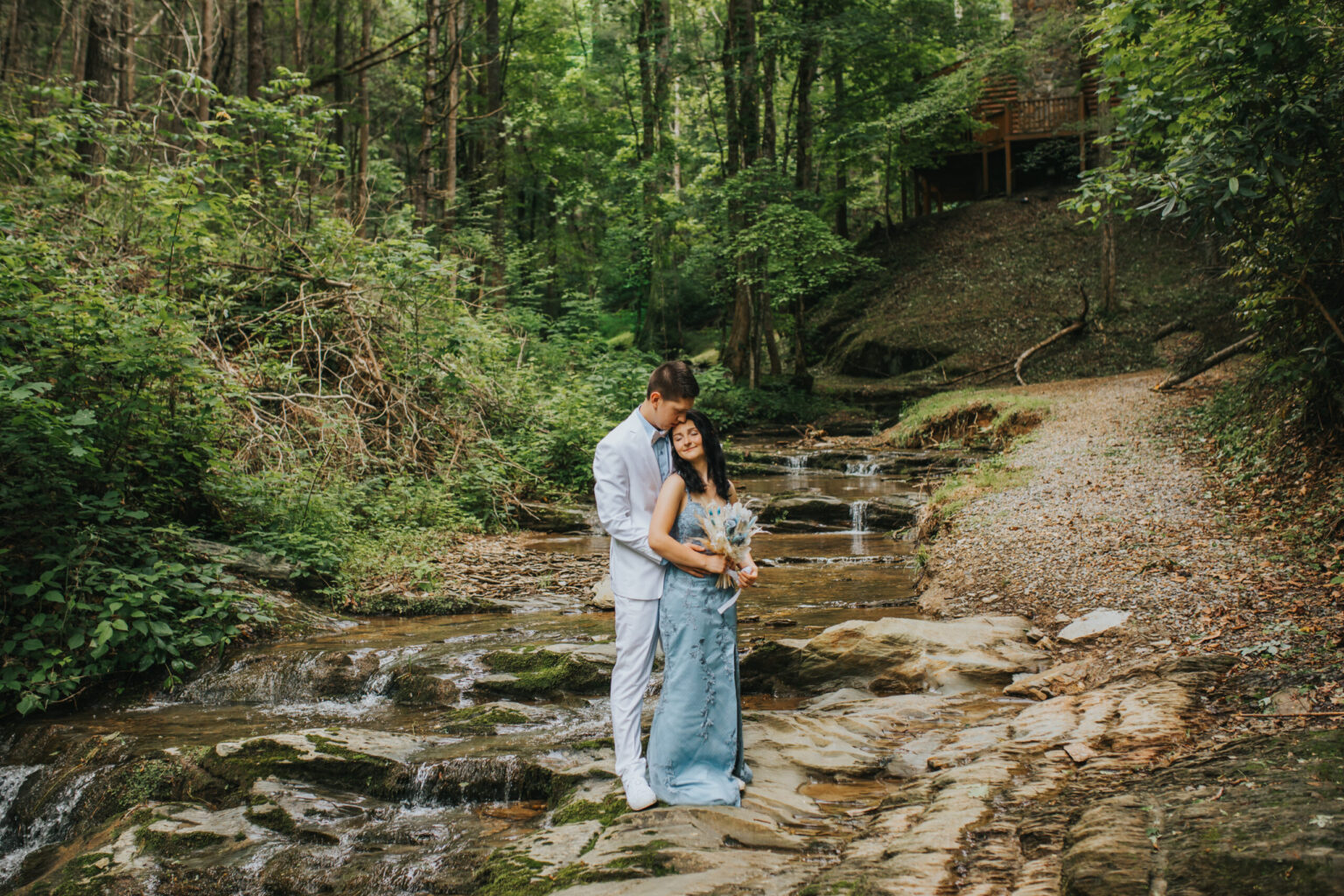 Elopement Wedding A couple stands lovingly by a small, rocky stream in a lush forest. The groom is in a light-colored suit while the bride wears a pastel blue strapless dress and holds a bouquet. They are eloping, surrounded by tall trees and greenery, with a rustic wooden structure visible in the background. Elopements Inc