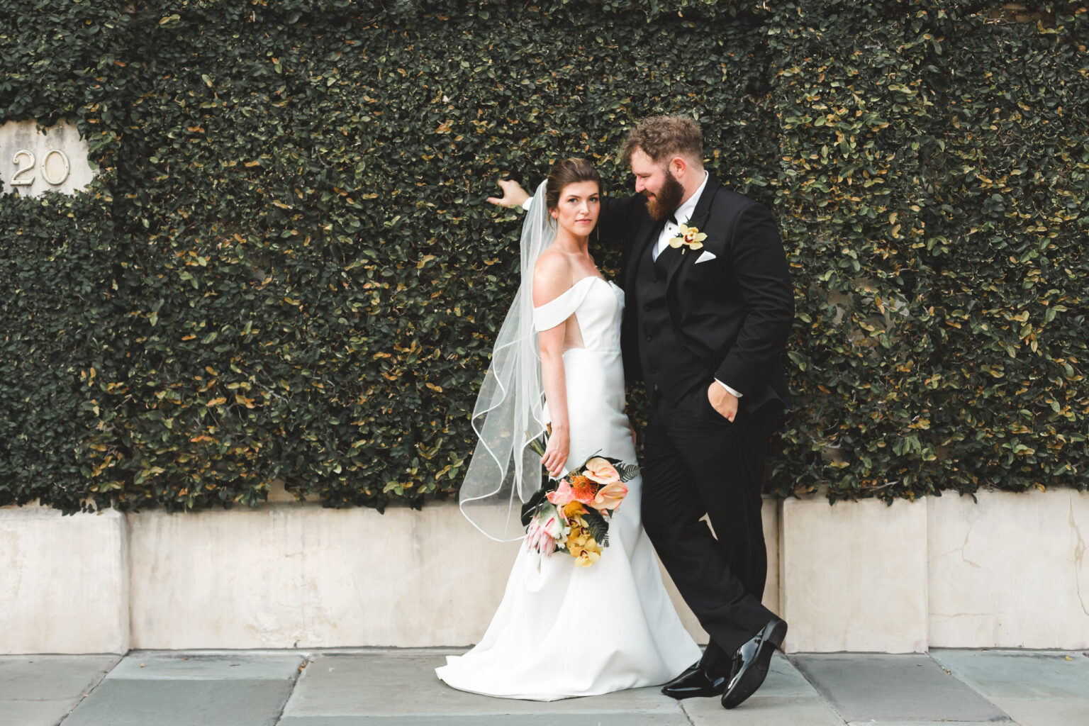 Elopement Wedding A bride in a white dress with a veil and a groom in a black suit pose against a lush ivy-covered wall. The bride holds a bouquet of flowers and smiles softly while the groom, leaning with one foot up, gazes at her. It's an intimate moment capturing the essence of their elopement amidst green ivy and stone. Elopements Inc