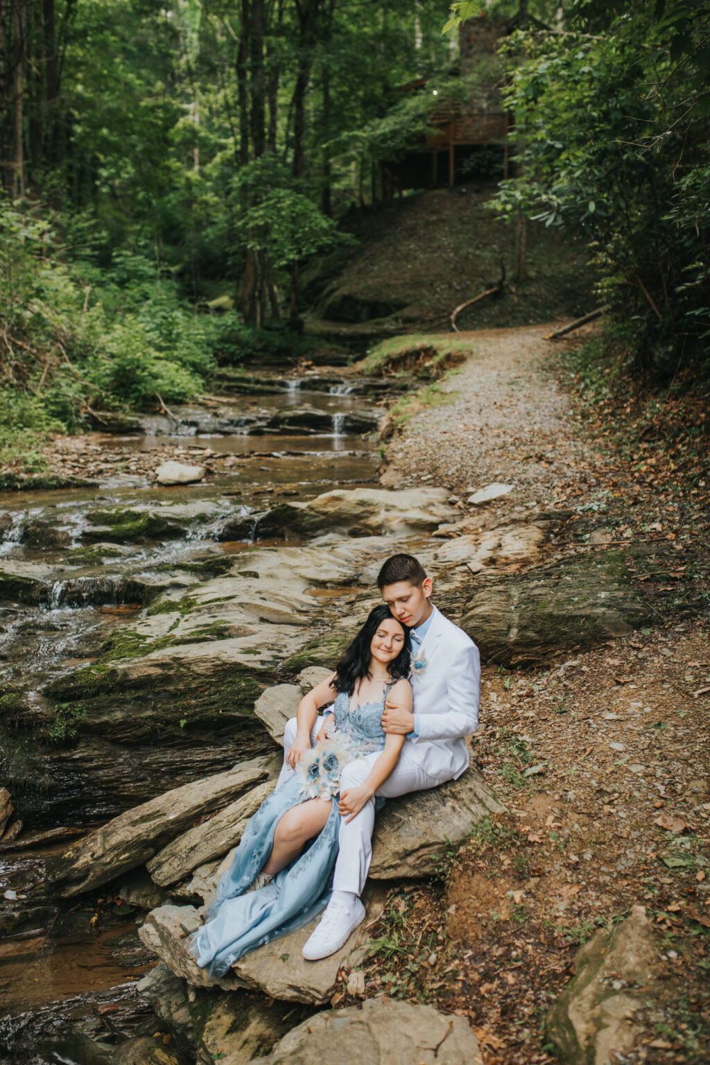 Elopement Wedding A man in a white suit and a woman in a light blue dress, having chosen to elope, sit on rocks beside a flowing stream in a forest. The woman leans against the man, resting her head on his shoulder, eyes closed. The background features dense green foliage, a dirt path, and a small wooden structure elevated on a slope. Elopements Inc