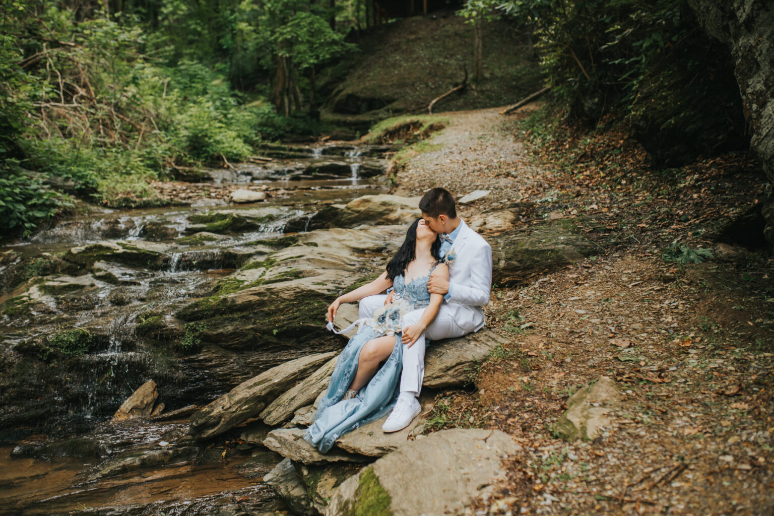 Elopement Wedding A couple sits on large rocks beside a tranquil forest stream, having just eloped. The woman, in a light blue dress, leans contentedly against the man, who is dressed in a white suit. They are surrounded by lush greenery and the rocky stream, creating a serene and romantic atmosphere. Elopements Inc