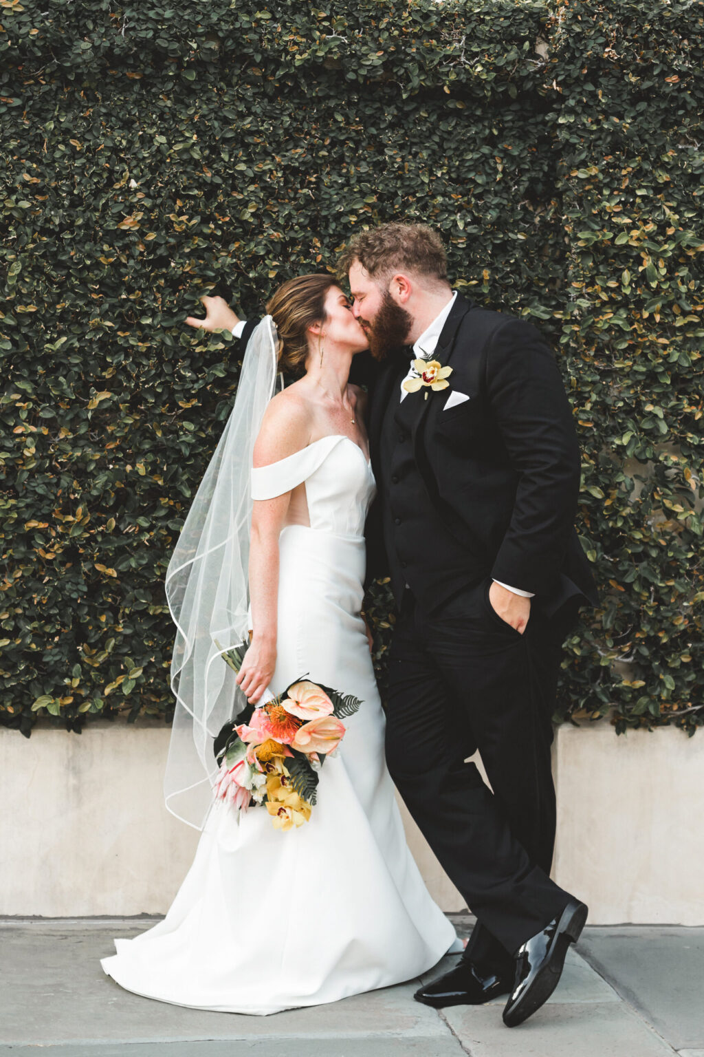 Elopement Wedding A bride and groom share a kiss in front of a lush green ivy wall, celebrating their intimate elopement. The bride, wearing a white strapless dress with a long veil, holds a bouquet of mixed flowers. The groom, dressed in a black tuxedo with a boutonniere that matches the bouquet, gently holds her waist while leaning against the wall. Elopements Inc