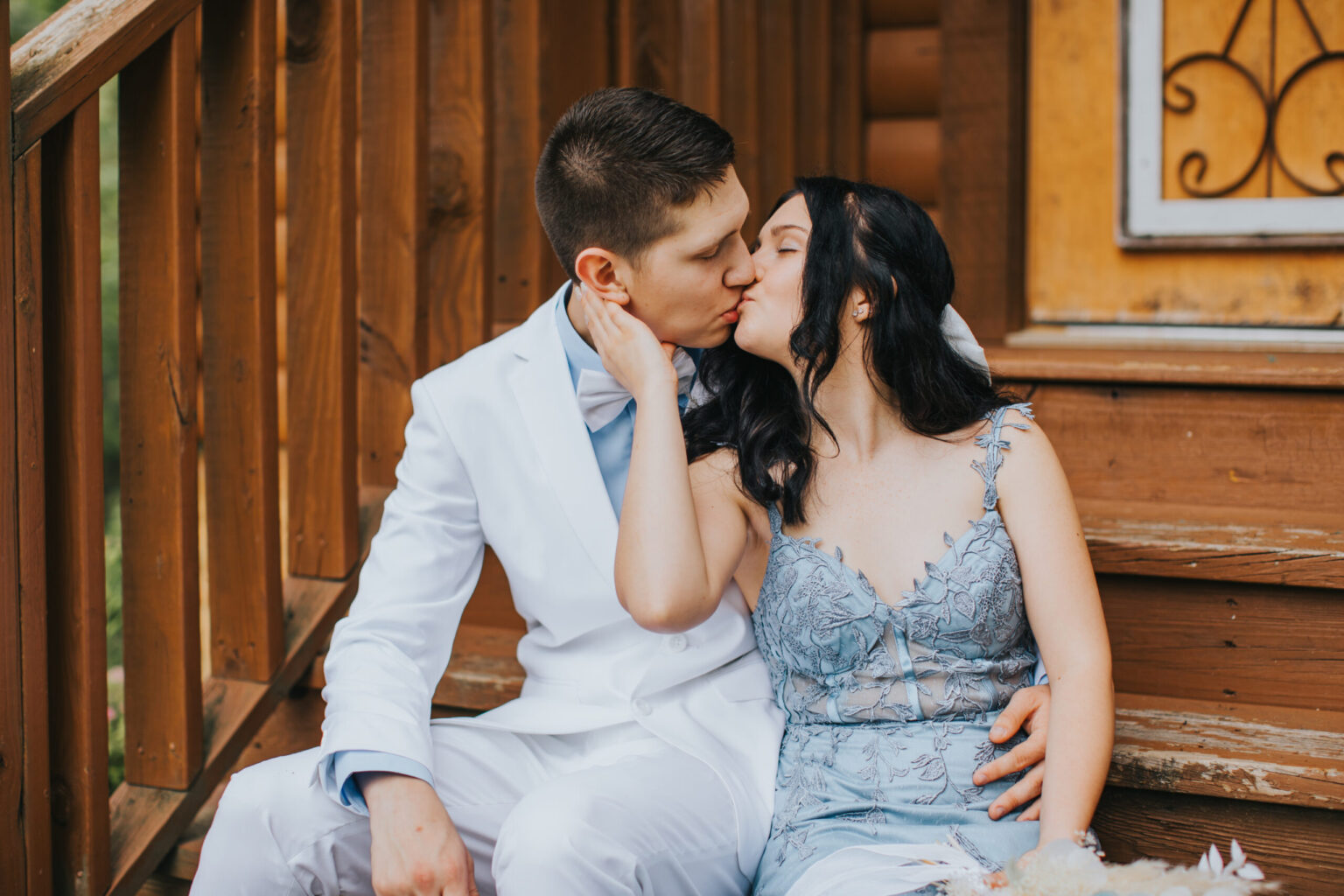 Elopement Wedding A couple sits on wooden steps, sharing a kiss during their intimate elopement. The person on the left is wearing a white suit with a bow tie, while the person on the right is in a light blue dress with lace details. They lean into each other, framed by part of a wooden structure and an iron decorative element. Elopements Inc