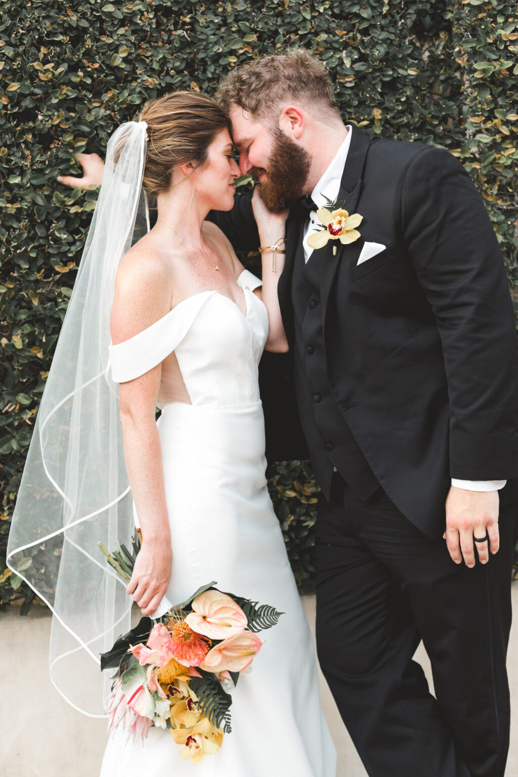 Elopement Wedding A bride and groom, having chosen to elope, lean in for an intimate moment in front of a greenery-covered wall. The bride, in an off-shoulder white gown and veil, holds a bouquet of tropical flowers. The groom, in a black suit with a boutonnière, touches his forehead to hers, both smiling gently. Elopements Inc