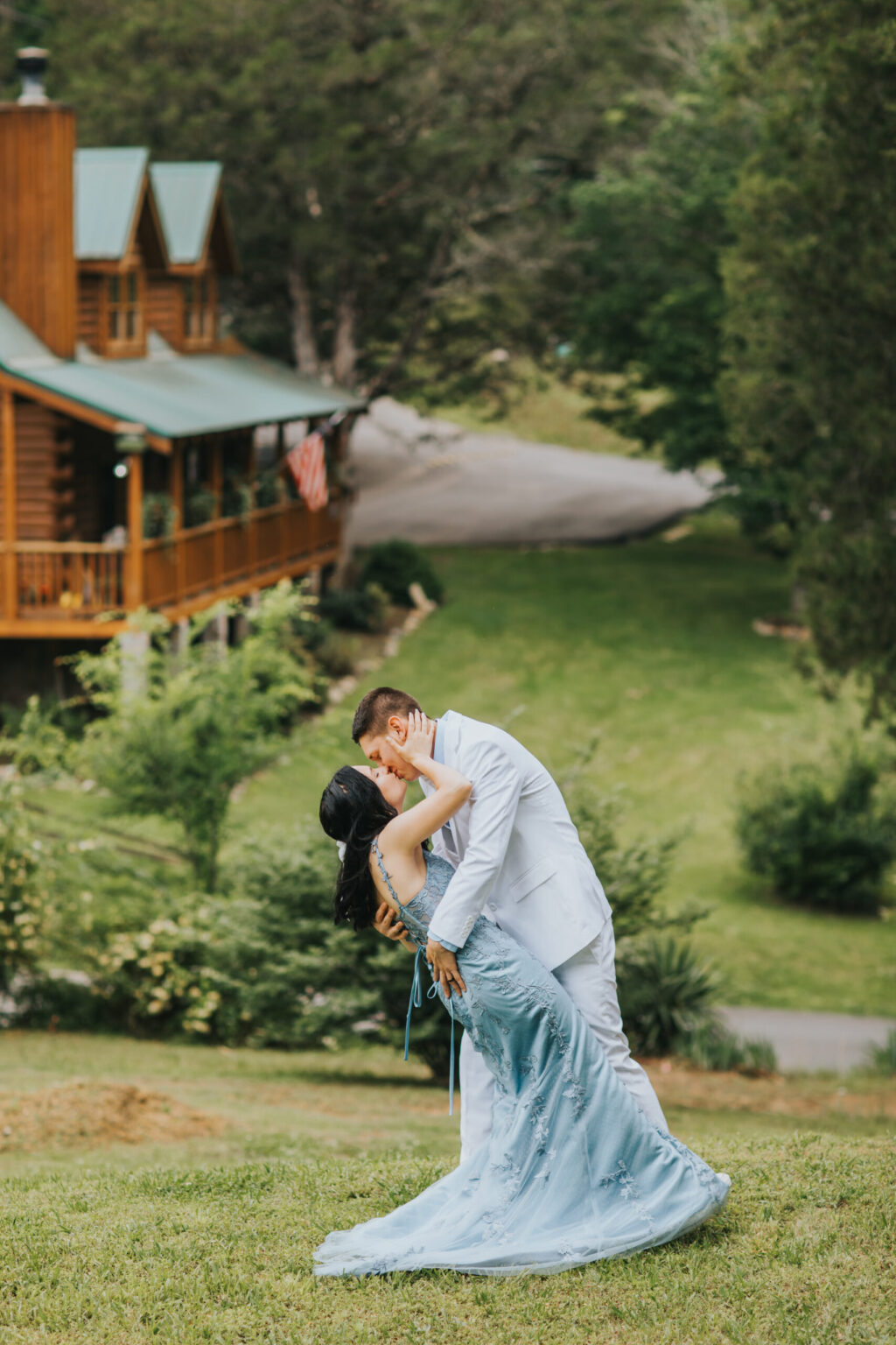 Elopement Wedding A couple, seemingly eloping, dressed in formal attire share a romantic dance in front of a quaint log cabin with green rooftops, surrounded by lush greenery. The man, in a light gray suit, dips the woman, who is wearing a light blue dress, as they gaze into each other's eyes. Elopements Inc