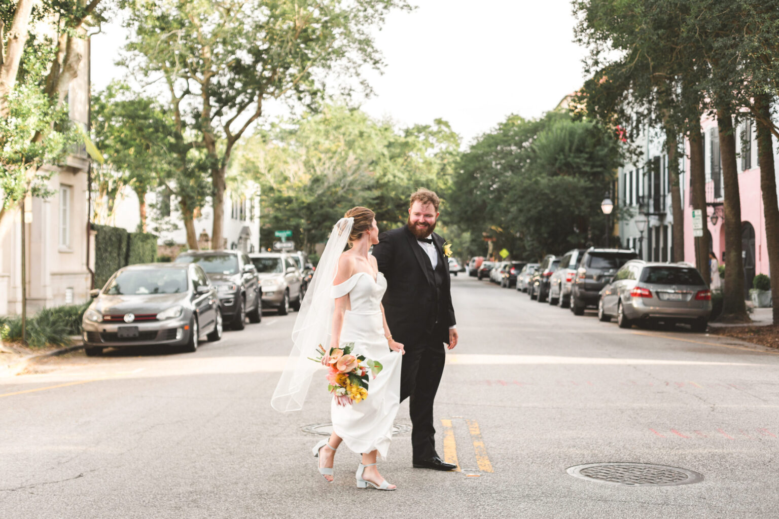 Elopement Wedding A bride in a white dress and veil walks with a groom in a black suit and bow tie across a street lined with parked cars and trees. The bride holds a bouquet of flowers, both smiling as they embrace their heartfelt elopement. The street is calm, with historic-looking buildings in the background. Elopements Inc