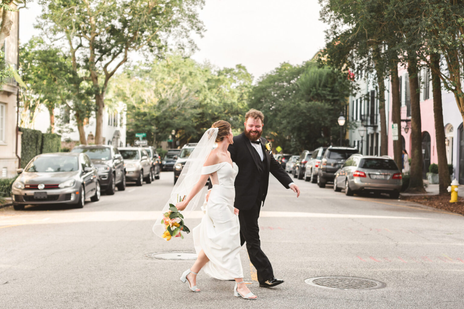 Elopement Wedding A bride and groom, having just eloped, hold hands as they walk joyfully across a street lined with parked cars and trees. The bride wears a white wedding dress with a veil and holds a bouquet of yellow and white flowers. The groom wears a black suit with a white shirt and black tie. Both are smiling and looking ahead. Elopements Inc