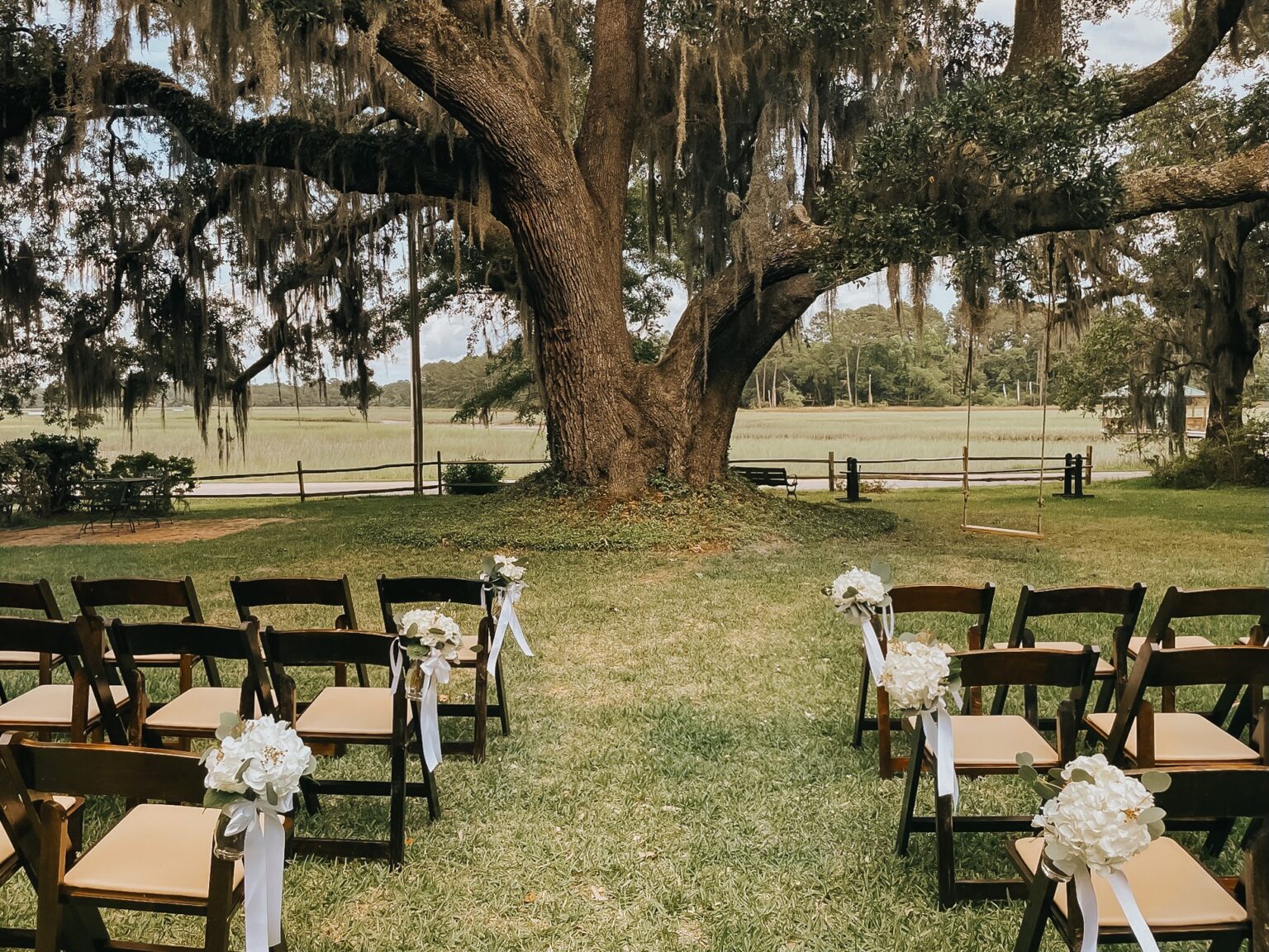 Elopement Wedding An outdoor wedding setup is depicted with rows of dark wooden chairs adorned with white flowers and ribbon, arranged on a grassy lawn. A large oak tree draped in Spanish moss stands at the center, with a scenic view of a coastal marshland extending into the background—a perfect setting for intimate elopements. Elopements Inc