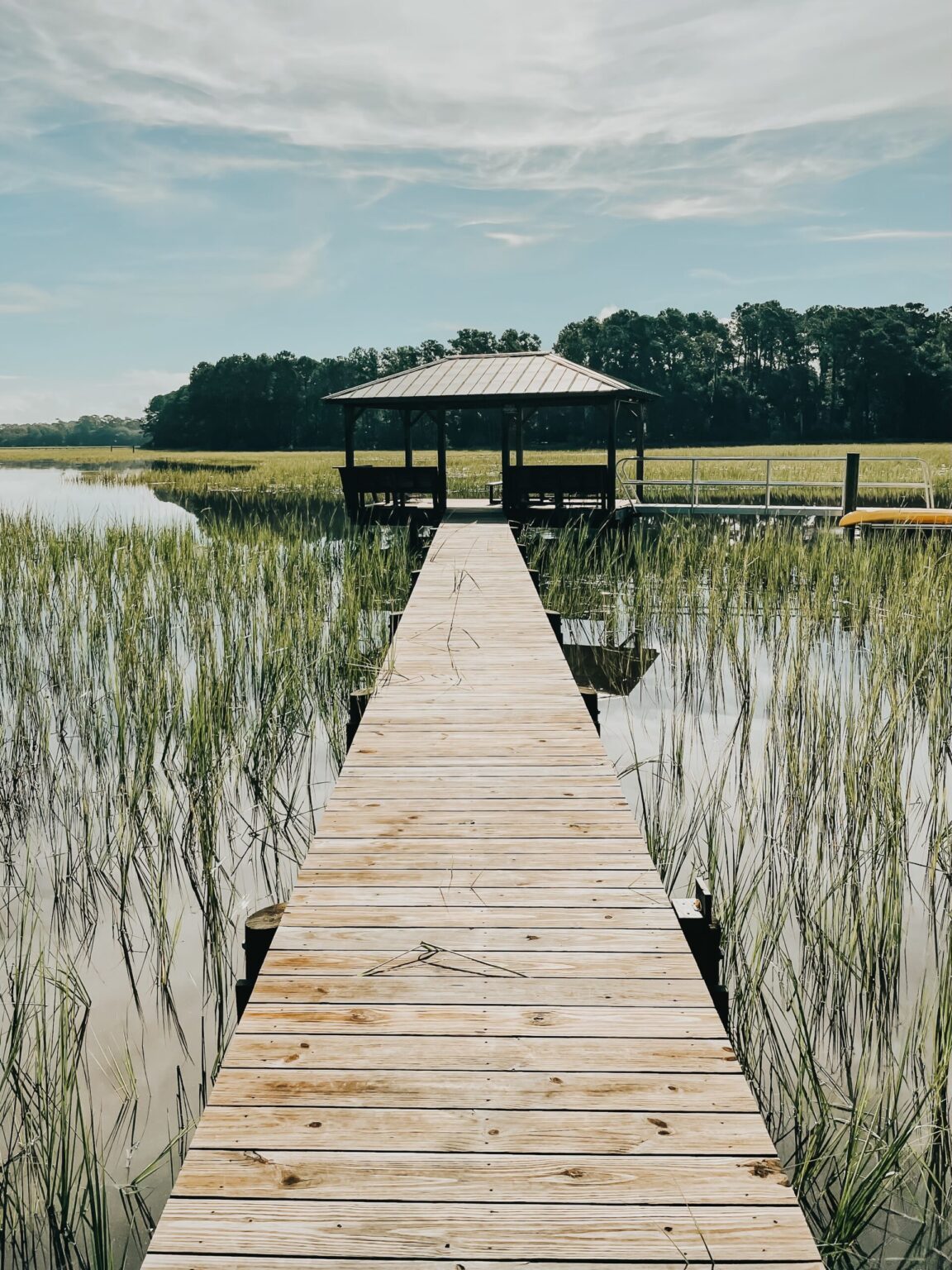 Elopement Wedding A wooden pier extends over marshy wetlands, leading to a covered gazebo. Tall grasses surround the water on both sides of the pier. A dense treeline is visible in the distance under a cloudy, yet bright sky. Elopements Inc