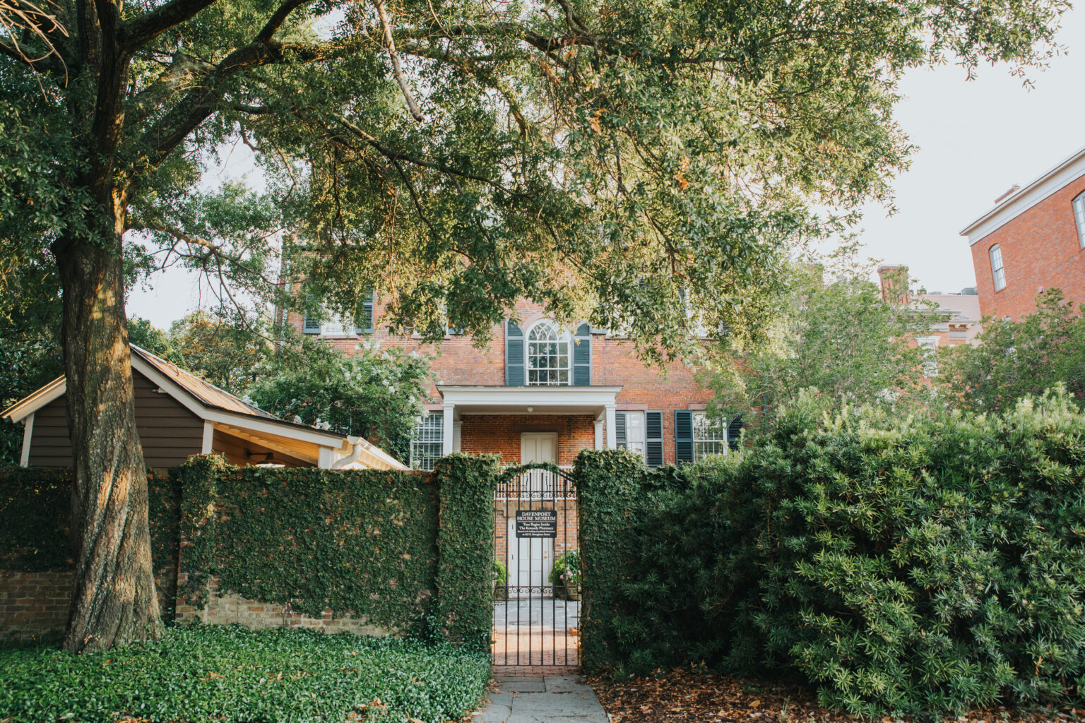 Elopement Wedding The image shows a brick two-story house partially obscured by a large tree and lush greenery. A black iron gate set in an ivy-covered wall marks the entrance. A brick path leads to the gate. To the left is a small wooden structure. The sunlit, serene scene exudes historic charm, perfect for intimate elopements. Elopements Inc