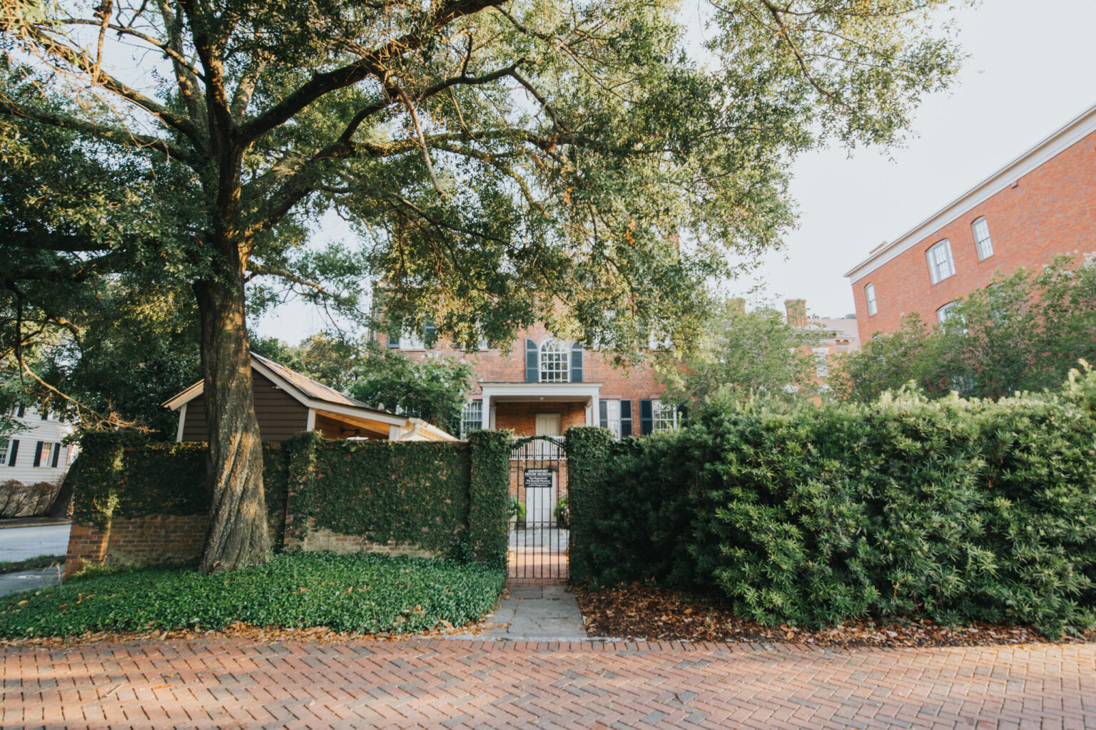 Elopement Wedding A serene residential scene showcasing a brick pathway leading to a gated entrance framed by dense greenery and a large mature tree. The gate, perfect for an intimate elopement, opens to a brick house partially visible behind the lush foliage on a sunny day, with another brick building in the background. Elopements Inc