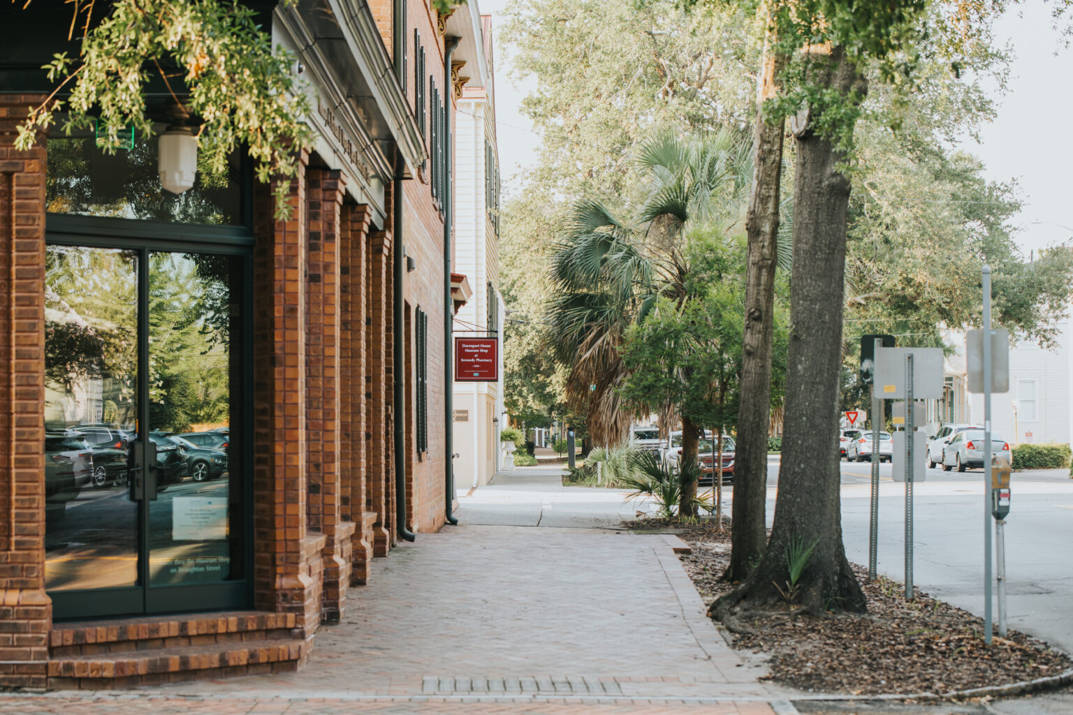 Elopement Wedding A quiet, tree-lined street with a brick-paved sidewalk and red brick buildings sets the perfect backdrop for an intimate elopement. A glass door entry reflecting a nearby car, accompanied by a red sign above. To the right, traffic light poles and an intersection with cars parked in the background complete the scene. Elopements Inc