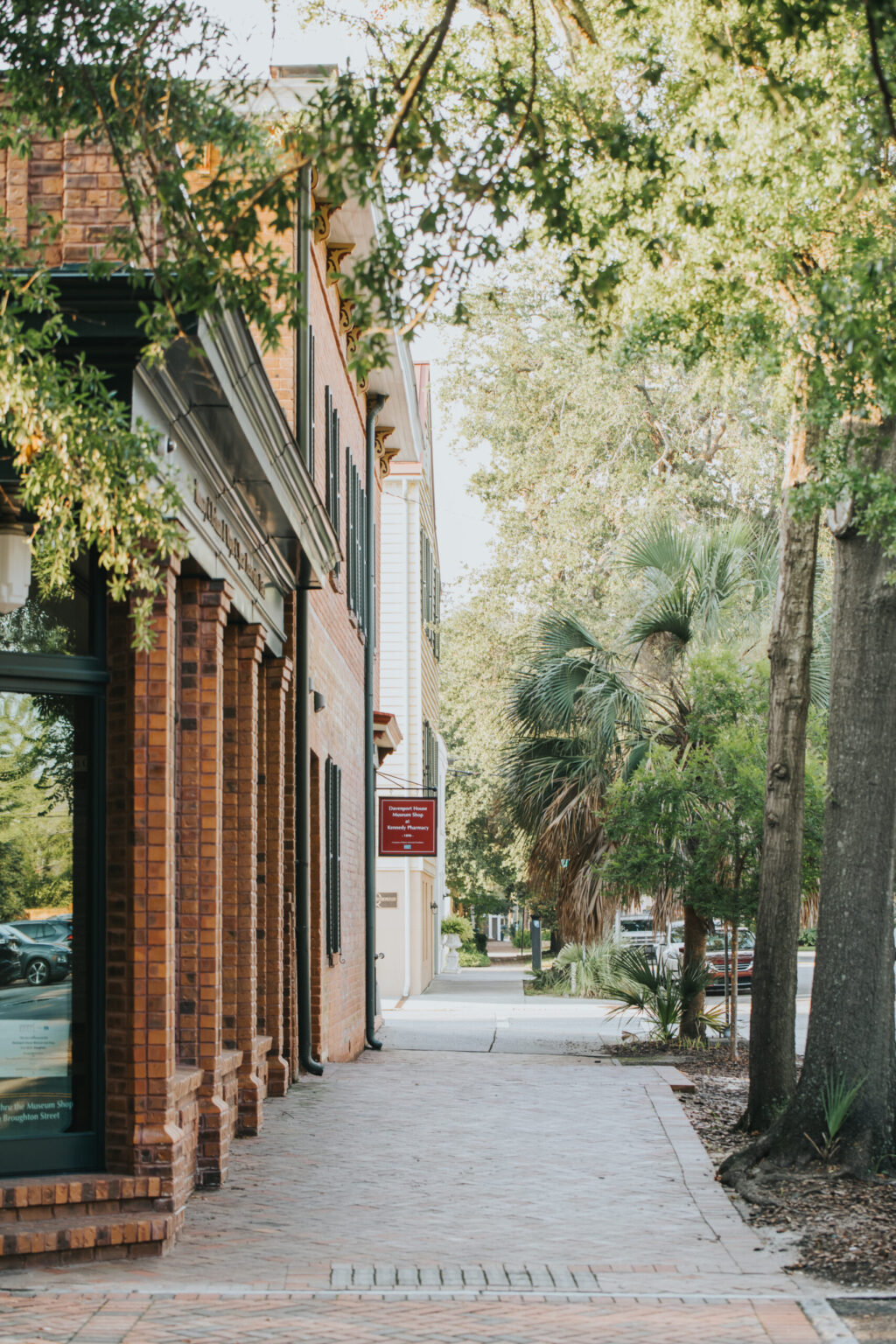 Elopement Wedding A quiet street corner features a brick building with black shutters on the windows. A red sign hangs near the building's entrance. The sidewalk, often chosen for intimate elopements, is shaded by trees, including a tall palm tree. The scene is bathed in natural light, giving it a serene and inviting atmosphere. Elopements Inc