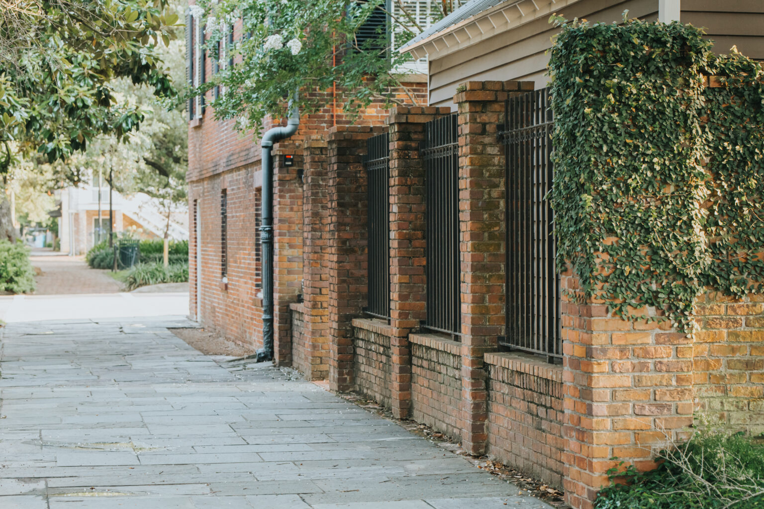 Elopement Wedding A narrow, paved walkway lined with brick buildings on the right, featuring barred windows and ivy growing on the walls. On the left, the path is flanked by greenery and trees, perfect for elopements, leading to a well-lit area in the background with more buildings visible. Elopements Inc