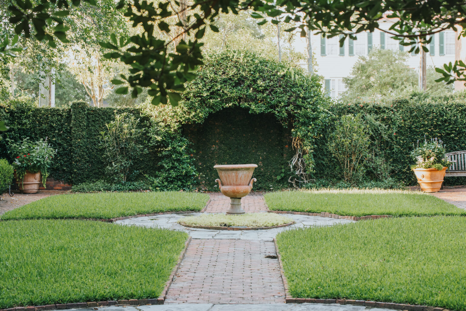 Elopement Wedding A well-manicured garden features trimmed green hedges and symmetrical grassy patches divided by brick pathways. In the center, a large decorative stone planter sits atop a circular base. Potted plants and a wooden bench on the right add charm, making it a picturesque spot for an elopement. Sunlight filters through the leafy trees above. Elopements Inc