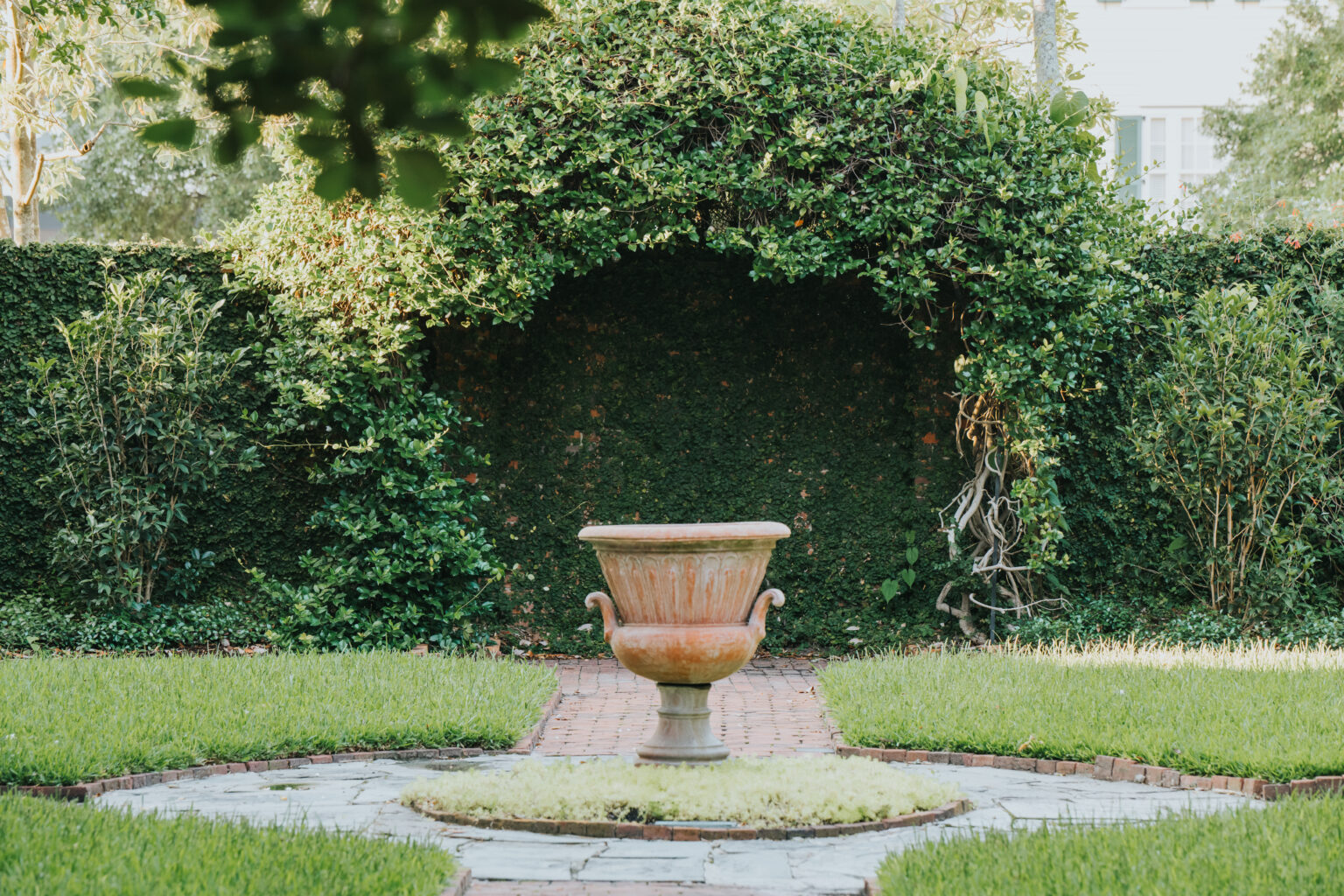 Elopement Wedding A serene garden, perfect for intimate elopements, features a large, empty terracotta planter as its focal point. The planter rests on a symmetrical stone pathway that leads to it. Surrounding the area are lush green hedges and leafy trees, creating a private and tranquil atmosphere. Brick accents add rustic charm to the setting. Elopements Inc