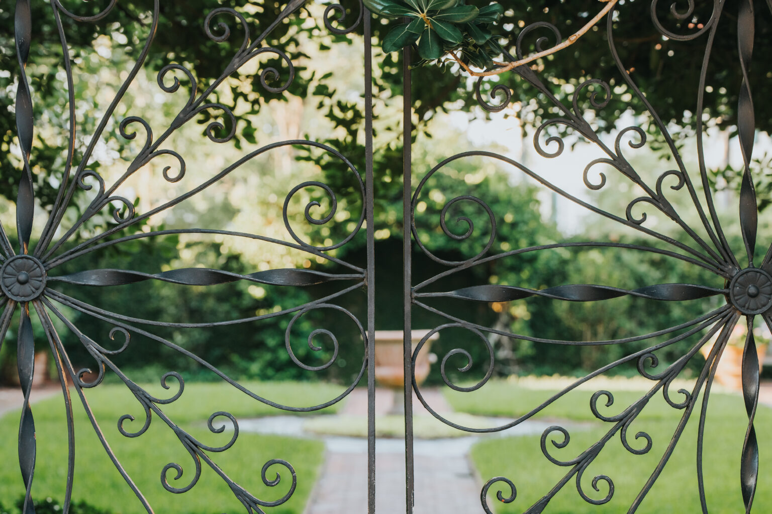 Elopement Wedding A wrought iron gate with intricate floral patterns is partially open, revealing a lush garden with a grassy lawn, a brick pathway, and a wooden bench in the background. Perfect for intimate elopements, the scene is framed by green foliage and dappled sunlight, creating a serene and inviting atmosphere. Elopements Inc