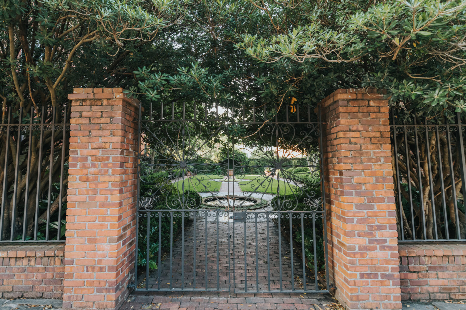 Elopement Wedding A wrought iron gate flanked by red brick pillars opens to a cobblestone path leading to a white stone fountain. The path, perfect for an elopement, is surrounded by lush greenery and tree branches that arch overhead. Sunlight filters through the foliage, creating a serene, inviting garden ambiance. Elopements Inc