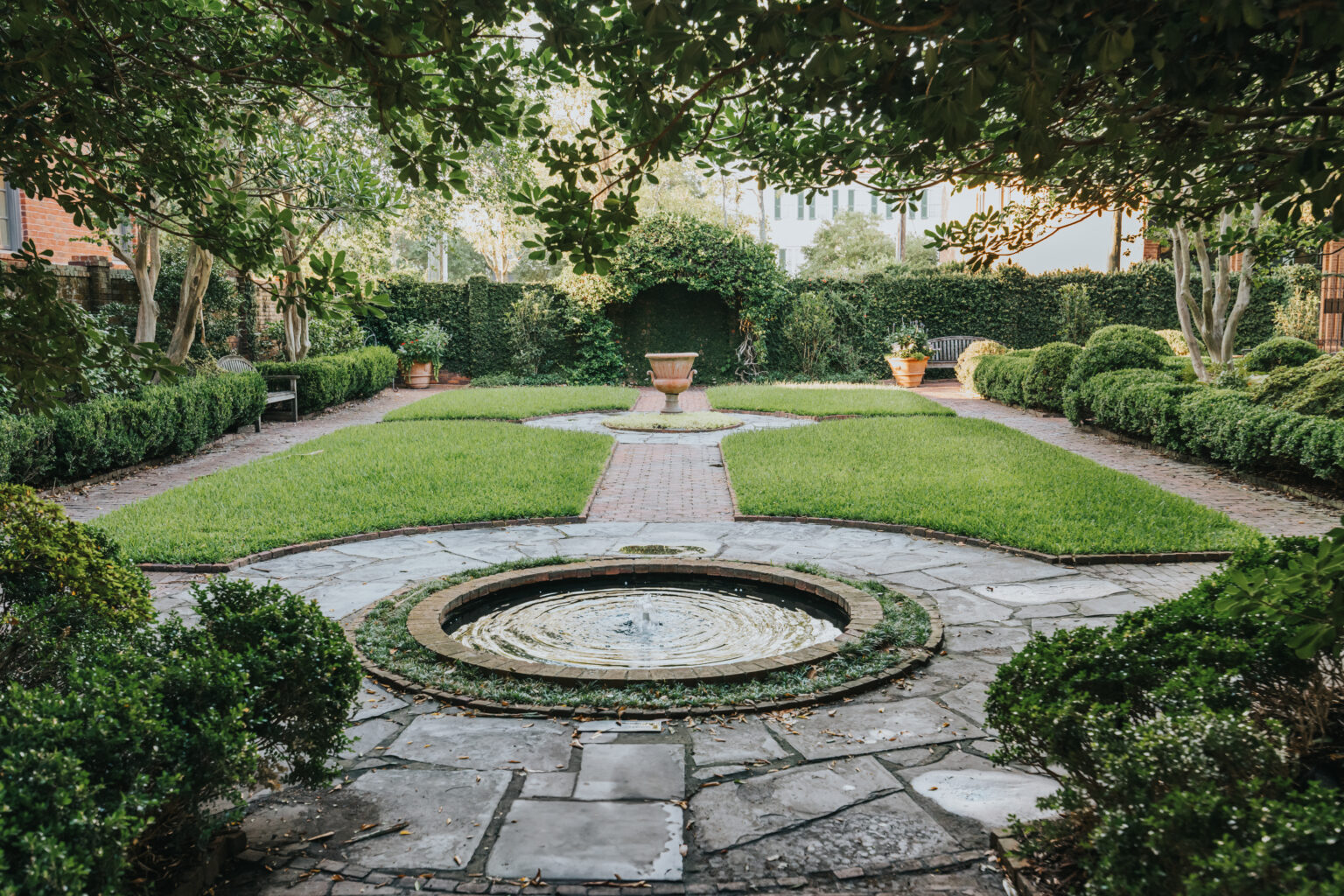 Elopement Wedding A serene garden features a circular fountain with rippling water in the foreground, surrounded by a stone pathway. Manicured green lawns and symmetrical hedges create a pathway leading to an ivy-covered archway, perfect for intimate elopements. Potted plants add to the lush, tranquil atmosphere. Elopements Inc