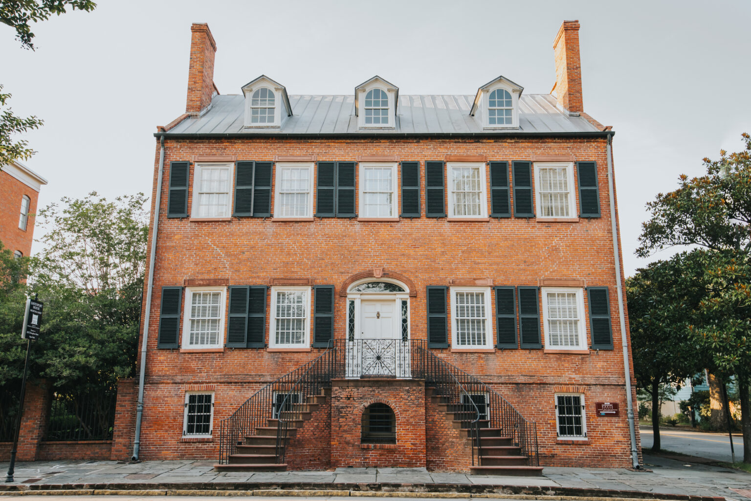 Elopement Wedding A historic, two-story red brick building with dark green shutters and a gray metal roof stands prominently. Perfect for intimate elopements, it features three dormer windows on the roof, a central white door with an arched transom, and a decorative wrought-iron railing on the front steps leading to the entrance. Trees frame both sides. Elopements Inc