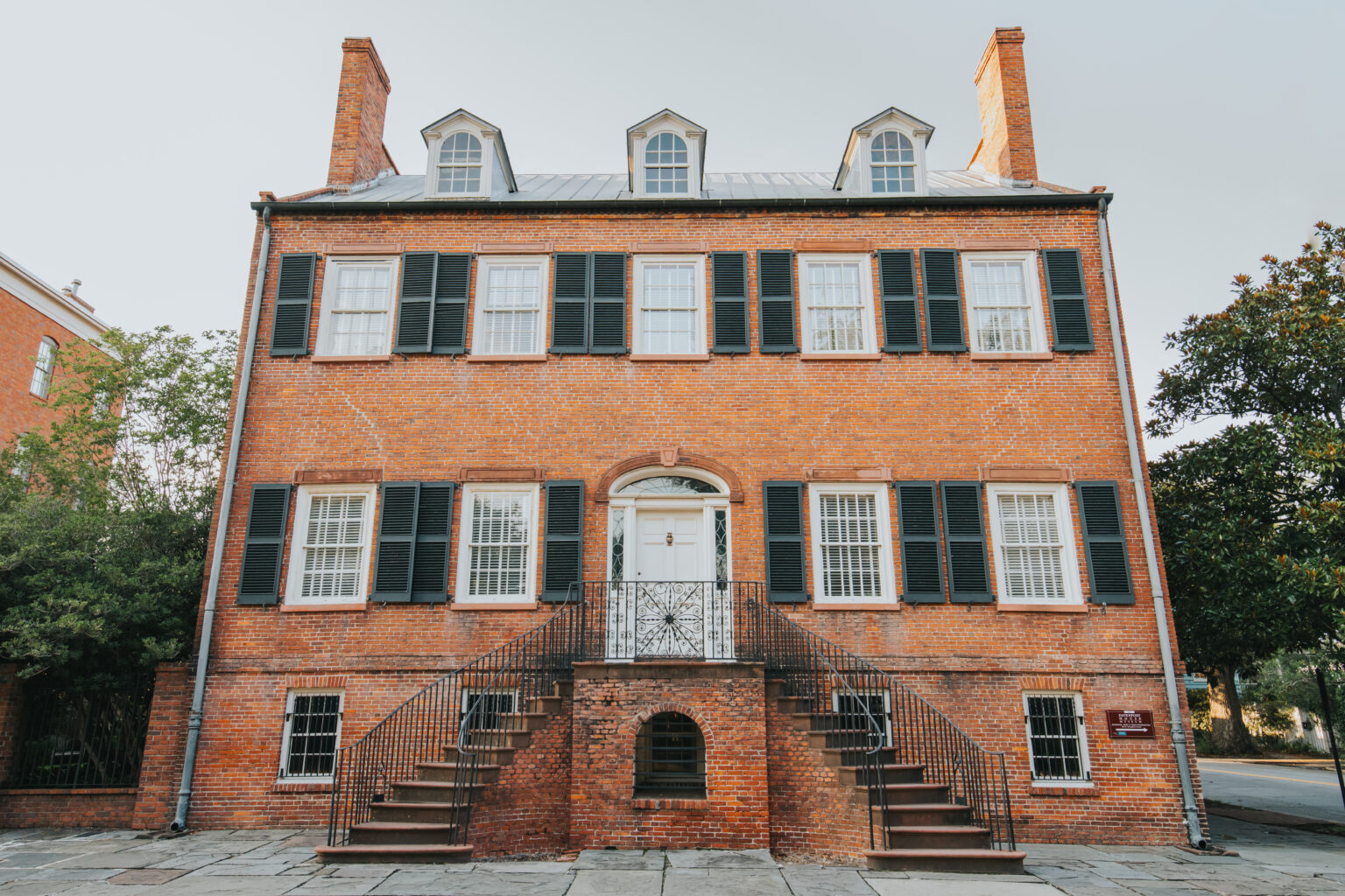 Elopement Wedding A large, historic brick building with three stories and a high-pitched roof featuring dormer windows sets a charming backdrop for couples eloping. The front has a symmetrical design with a central white door, flanked by black shutters on all windows. A double staircase leads to the entrance. Trees and adjacent buildings are visible. Elopements Inc