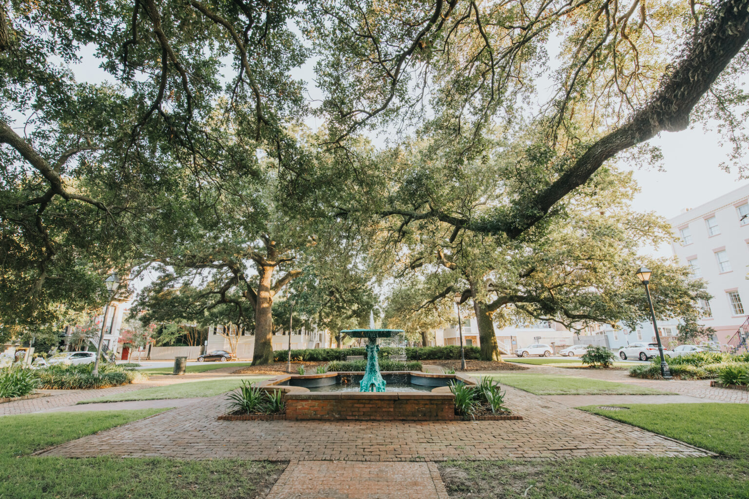 Elopement Wedding A picturesque park scene features a central fountain surrounded by brick pathways. Large, sprawling trees with abundant leaves provide shade, creating a serene atmosphere perfect for an intimate elopement. The background shows parked cars and buildings, while green lawns and manicured bushes add to the park's beauty. Elopements Inc