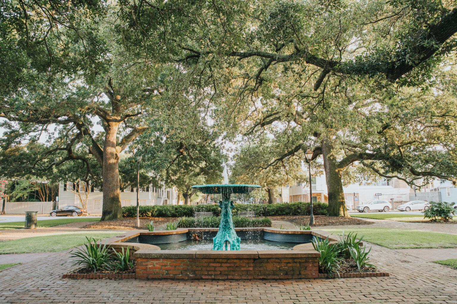 Elopement Wedding A tranquil park scene featuring a central water fountain surrounded by greenery, an ideal spot for couples eloping. The fountain has a three-tiered structure with blue-green water cascading down. Large oak trees with sprawling branches frame the background, and a brick pathway encircles the fountain. Elopements Inc