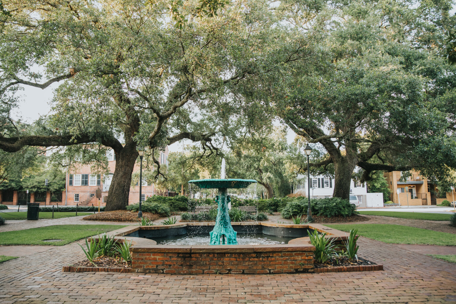 Elopement Wedding A serene outdoor scene perfect for elopements features a central fountain with a green, turquoise hue and water cascading down. The fountain sits in a small, square brick pool surrounded by red brick paving. Large, leafy trees arch overhead, creating a canopy, and residential buildings are visible in the background. Elopements Inc