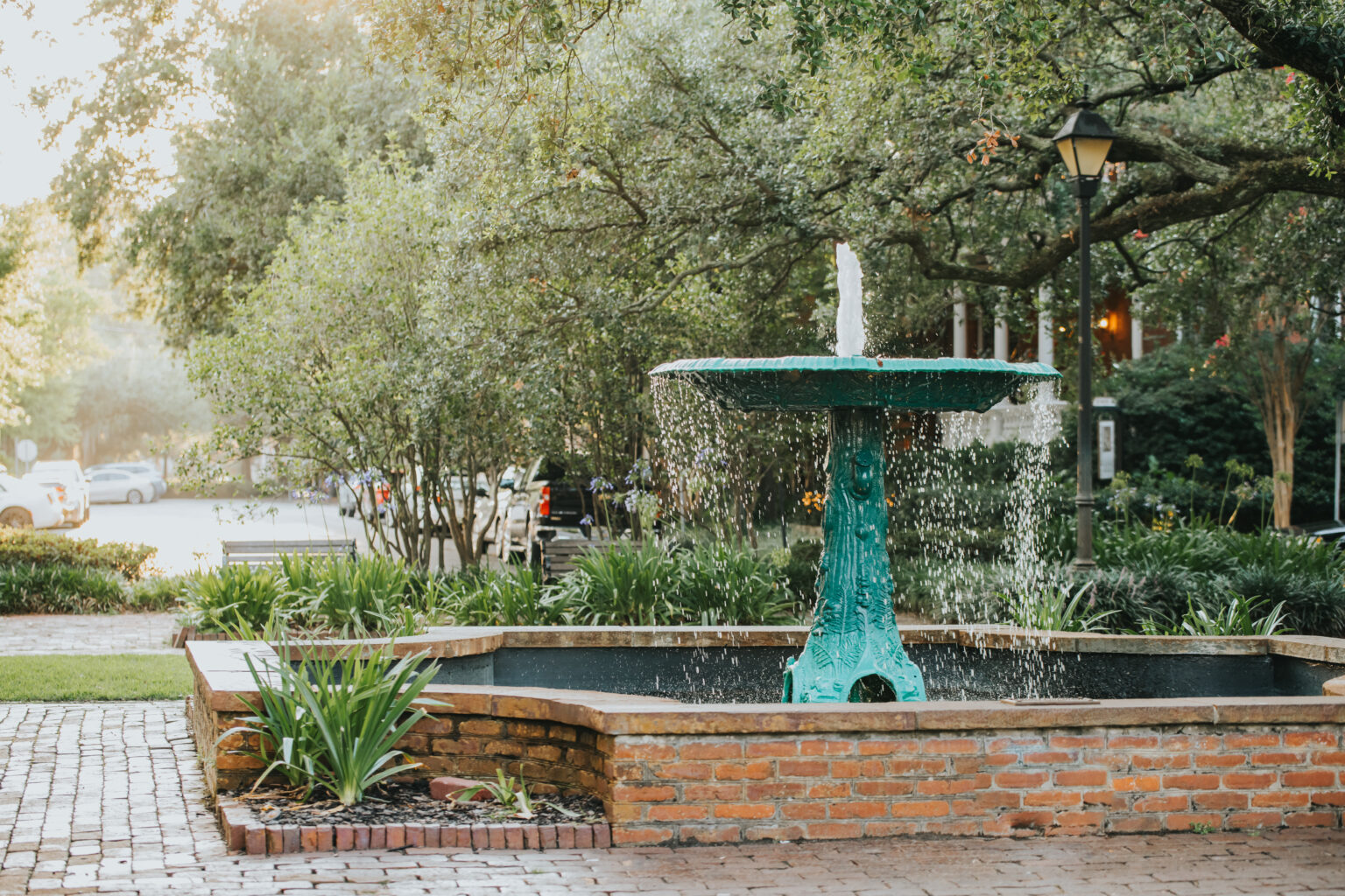 Elopement Wedding A tranquil scene of a green fountain in a small park surrounded by lush trees and plants sets the perfect backdrop for intimate elopements. Water cascades gently from the top tier of the fountain into a brick-lined basin, while a lamppost and some parked cars are bathed in the soft glow of early morning light. Elopements Inc