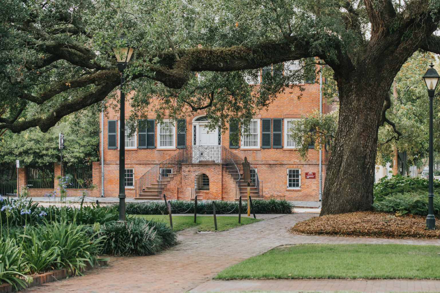 Elopement Wedding A historic red-brick building with dark shutters and a central white door framed by an ornate wrought-iron railing stands at the end of a tree-lined path. The large oak trees and lampposts enhance the old-world charm, perfect for an intimate elopement, with lush greenery and manicured landscaping completing the scenic setting. Elopements Inc