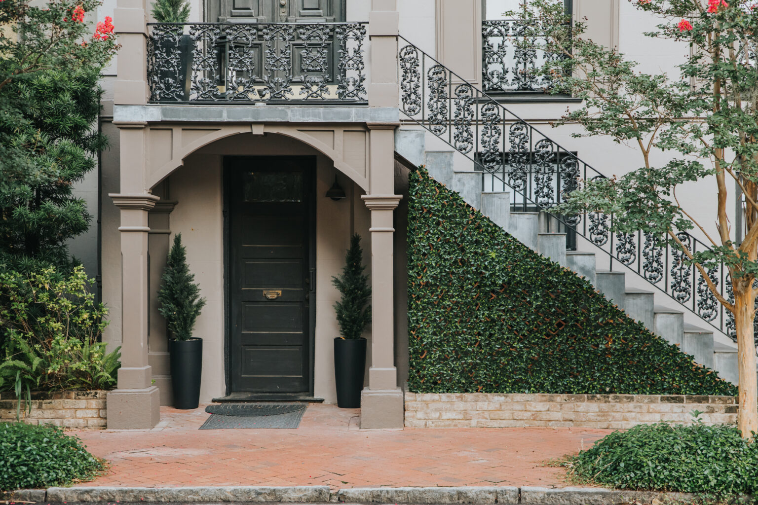 Elopement Wedding A black door is set in a light grey, arched entryway with decorative black iron railings on stairs to the right. Perfect for an intimate elopement, potted greenery flanks the door, and a sloped hedge extends beneath the staircase. The building’s façade features plants and a brick walkway in front. Elopements Inc