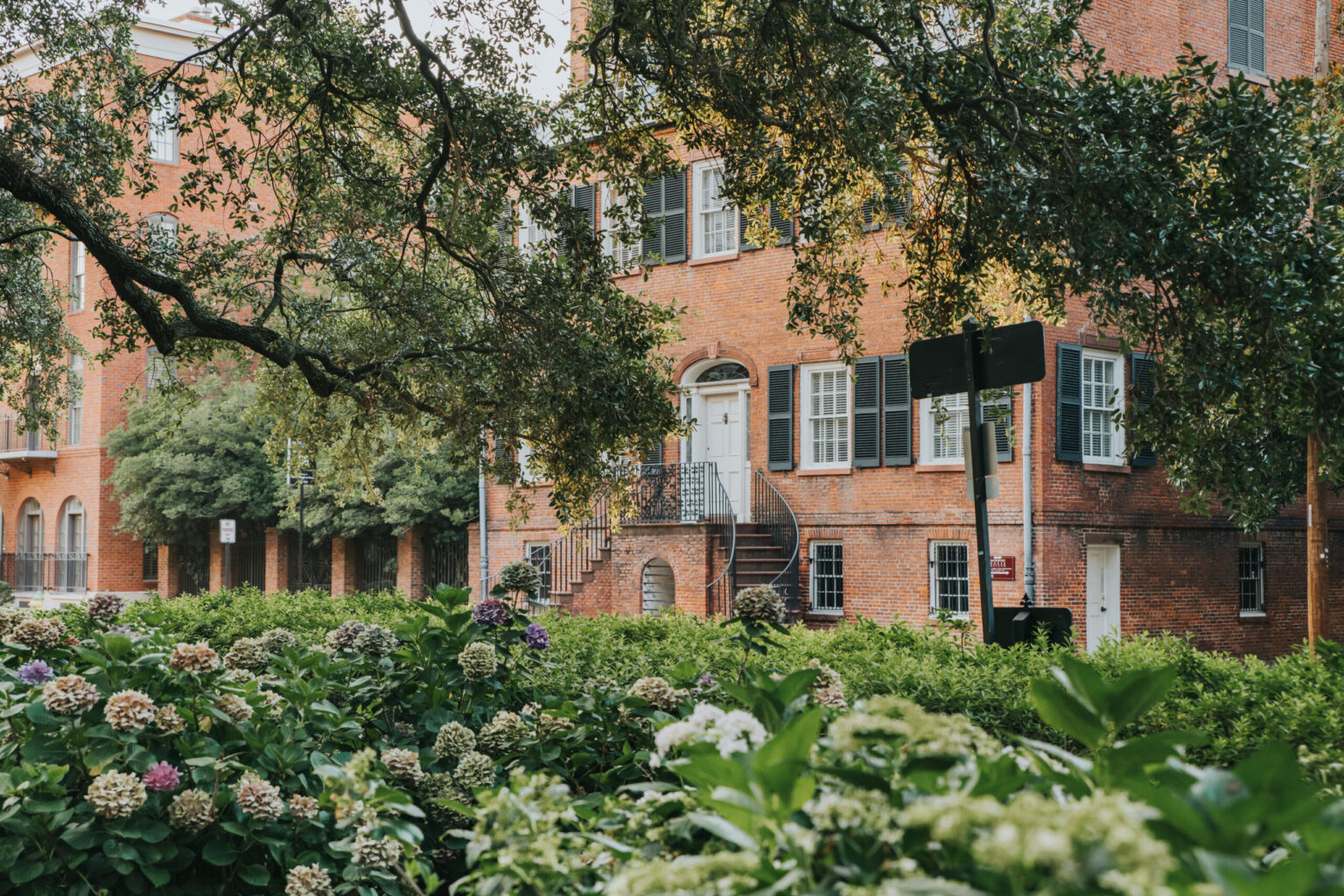 Elopement Wedding A stately brick building is partially obscured by lush greenery and blooming hydrangeas. A spiral staircase leads to a white door with large windows featuring shutters. An overhanging tree branch adds a picturesque touch, creating an idyllic backdrop perfect for intimate elopements in this charming urban setting. Elopements Inc