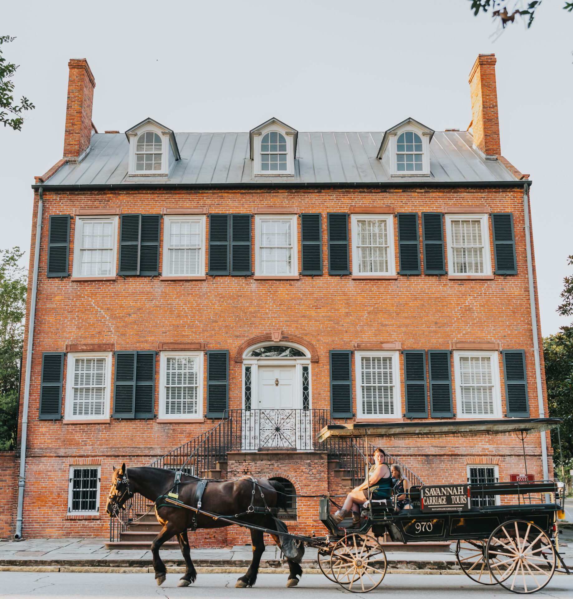 Elopement Wedding A historic red-brick building with black shutters and a gray roof features three dormer windows. A horse-drawn carriage labeled "Savannah Ghost Tours" waits nearby on the street. A person sits on the carriage seat, holding the reins of two black horses. Trees are visible on the edges of the image. Elopements Inc